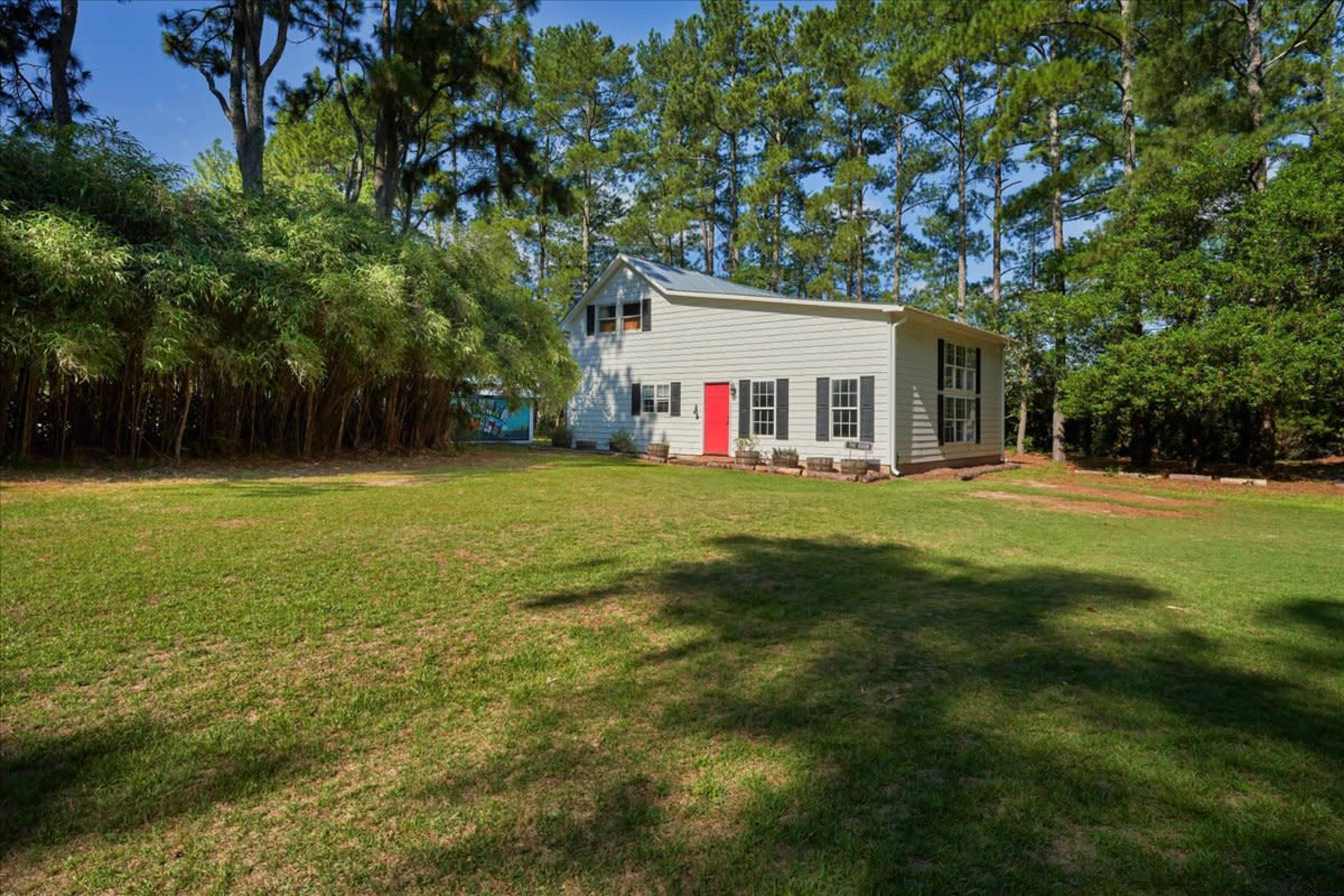 A two-story gray house with a red door is set in a grassy yard surrounded by tall trees and bamboo.