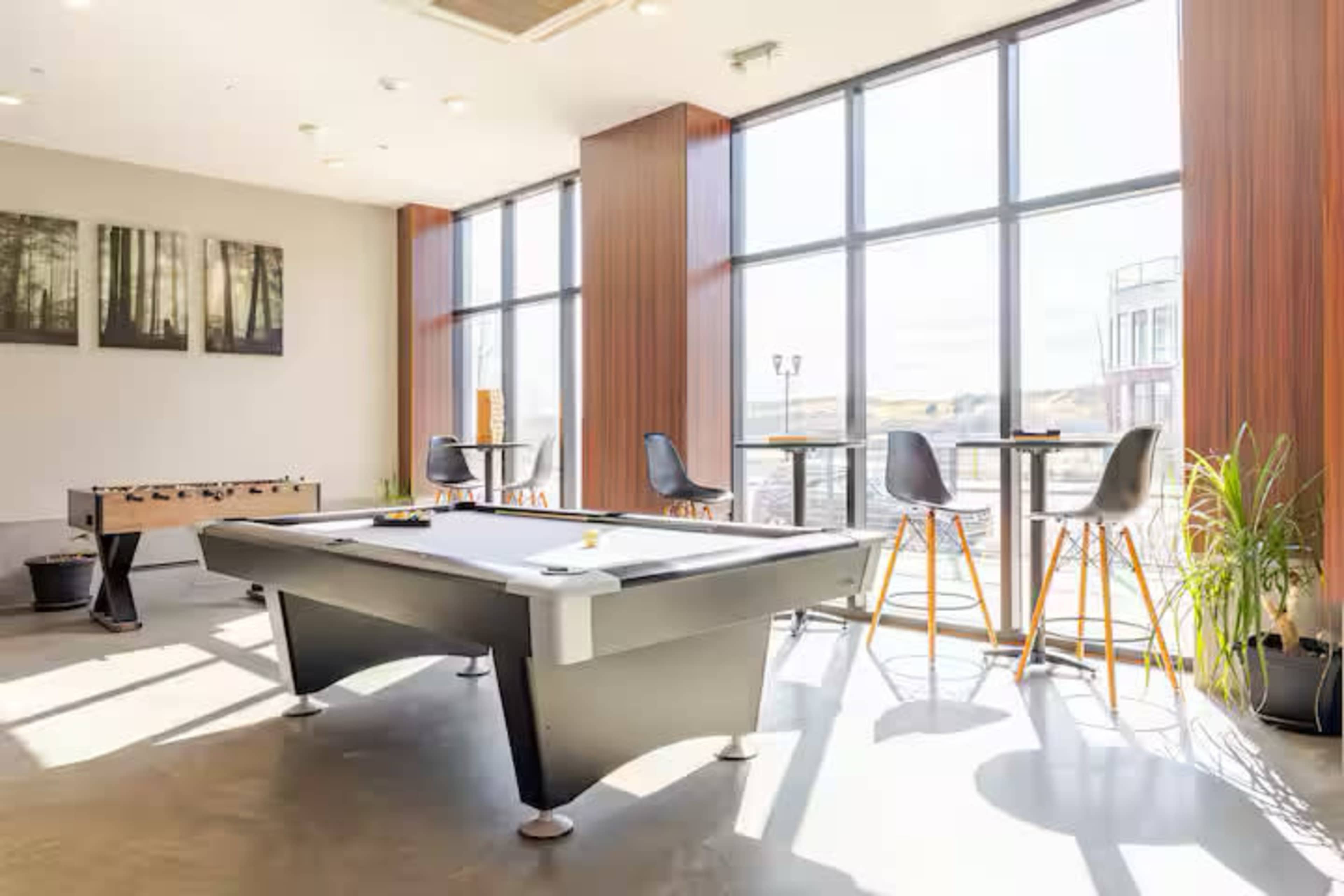 The image shows a bright recreational room featuring a pool table, foosball table, and bar seating near large windows.