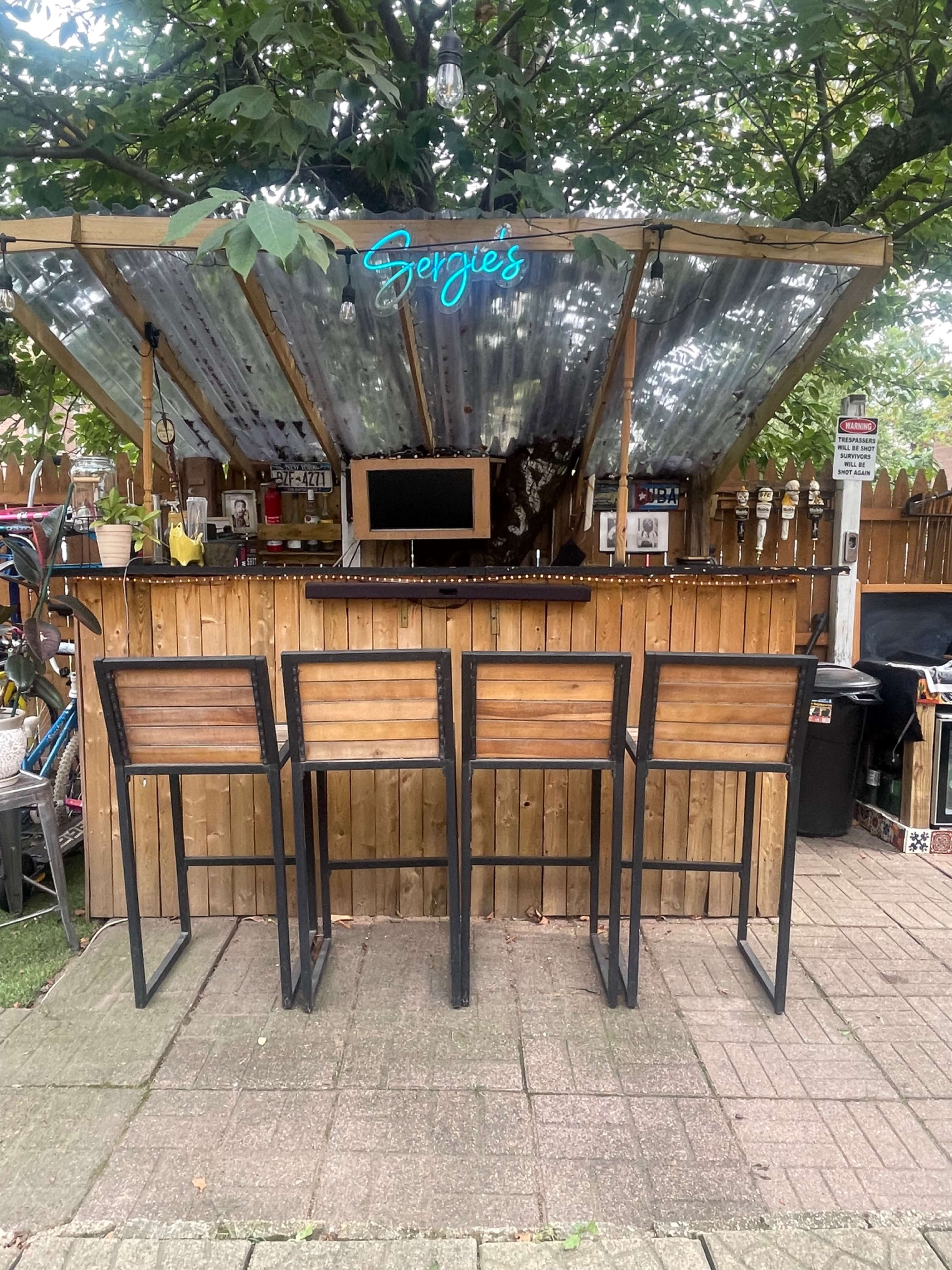 An outdoor bar with a wooden counter, four high stools, and a neon sign reading "Sergie's," under a shelter made from corrugated metal, surrounded by trees and garden decor.