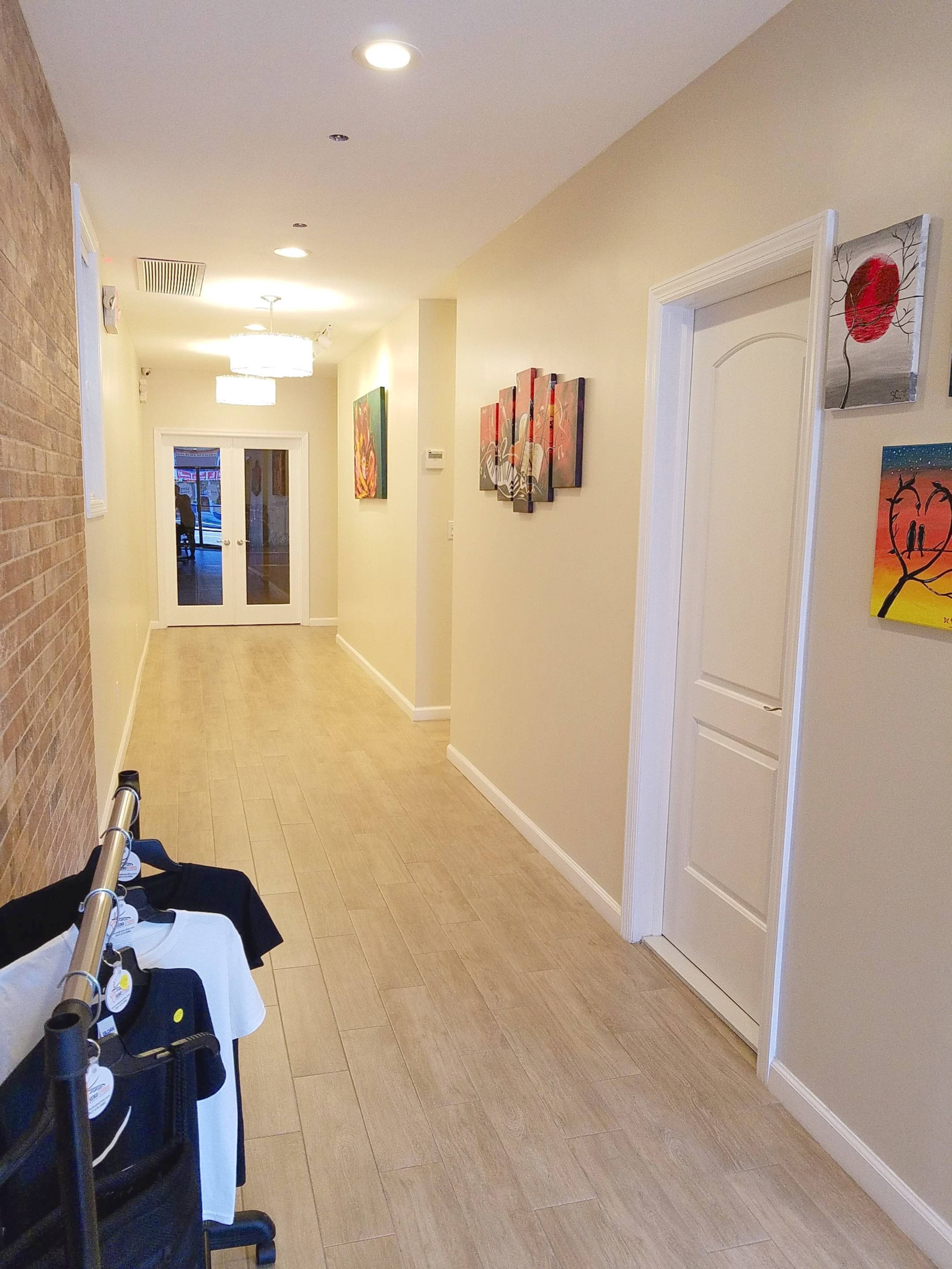 The image shows a well-lit hallway with a smooth tile floor, featuring artwork on the walls and a door at the end leading outside.