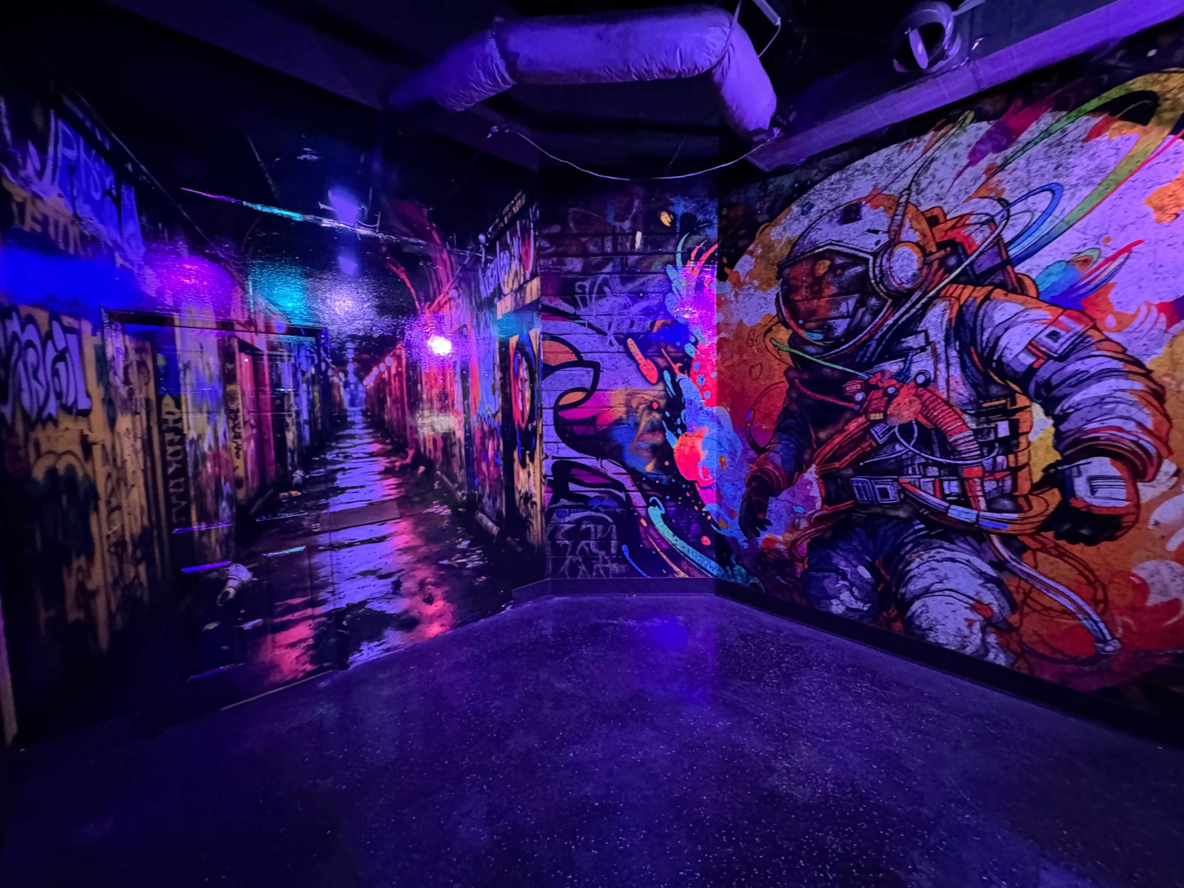 The image shows a vibrant mural featuring a colorful astronaut alongside a dark alley, illuminated by purple lights.