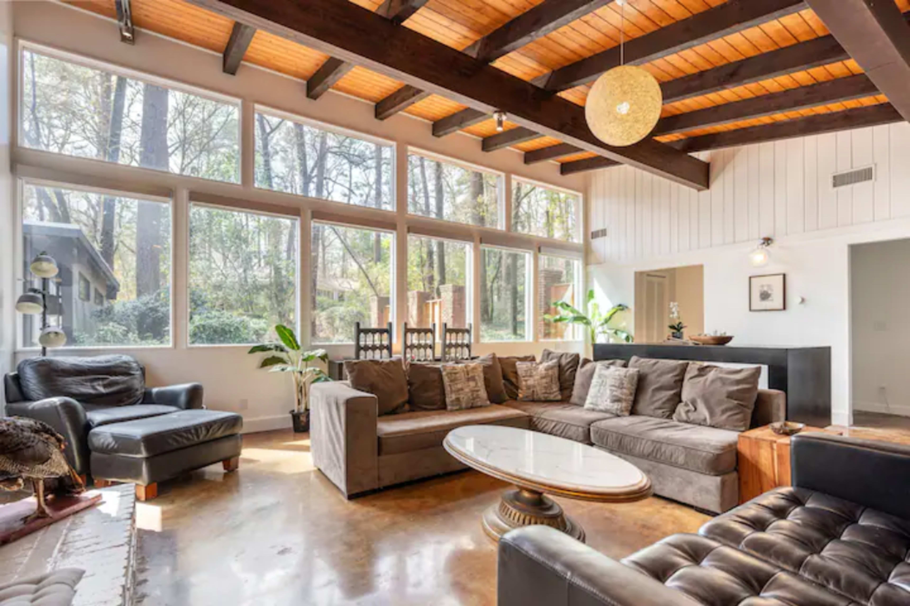 A modern living room with large windows allowing natural light, featuring a sectional sofa, a round coffee table, and wooden beams on the ceiling.