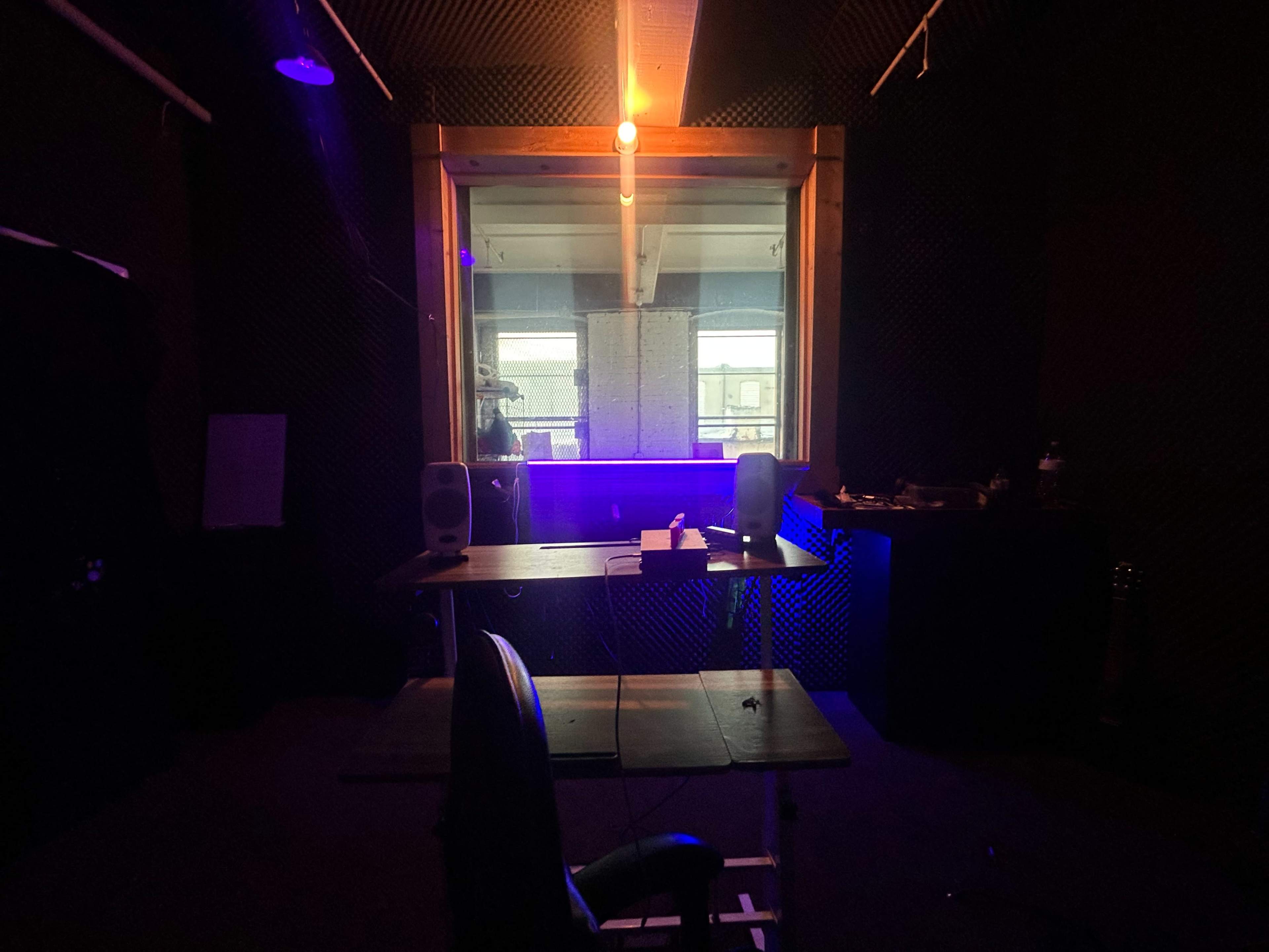 A dimly lit recording studio features a desk with speakers and a chair, facing a window that separates it from a brighter area.