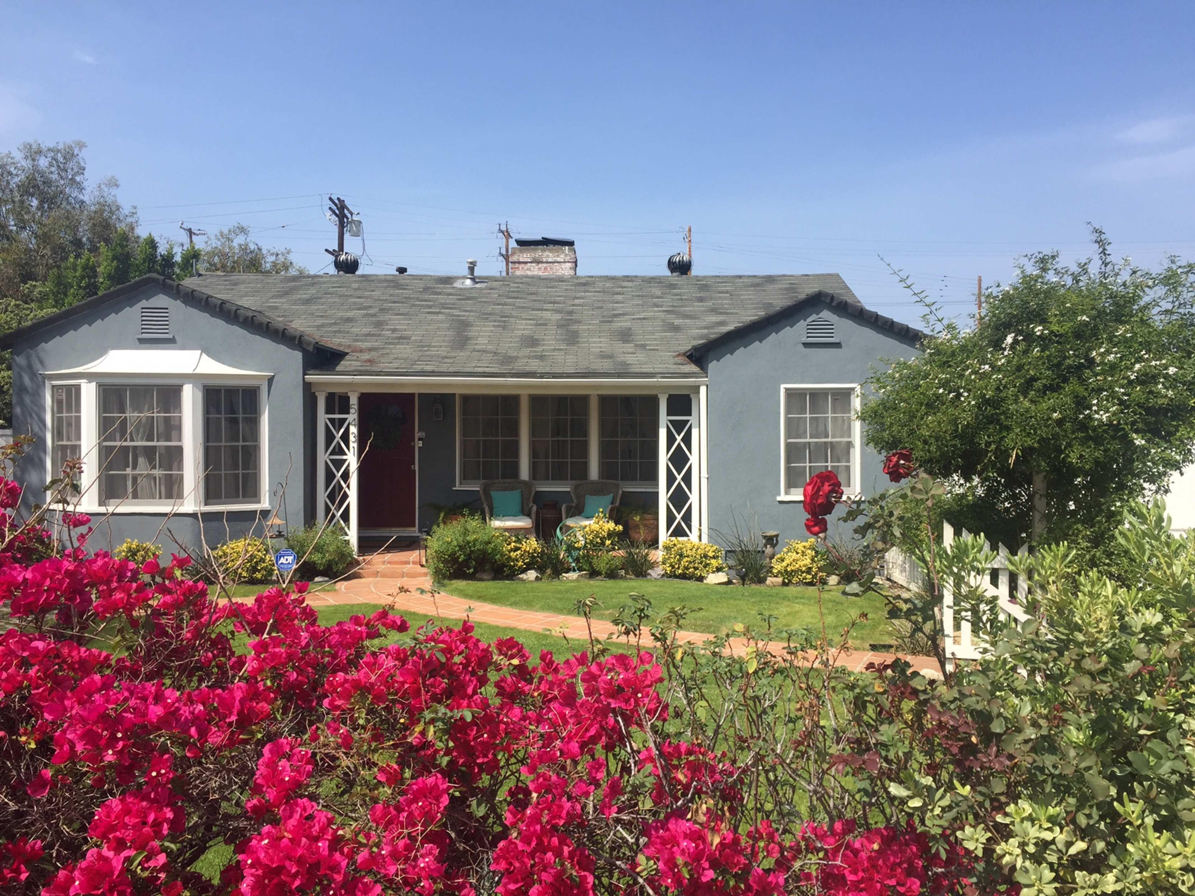 A light blue house with a shingled roof is surrounded by colorful flowers and features a red front door along with a small porch.