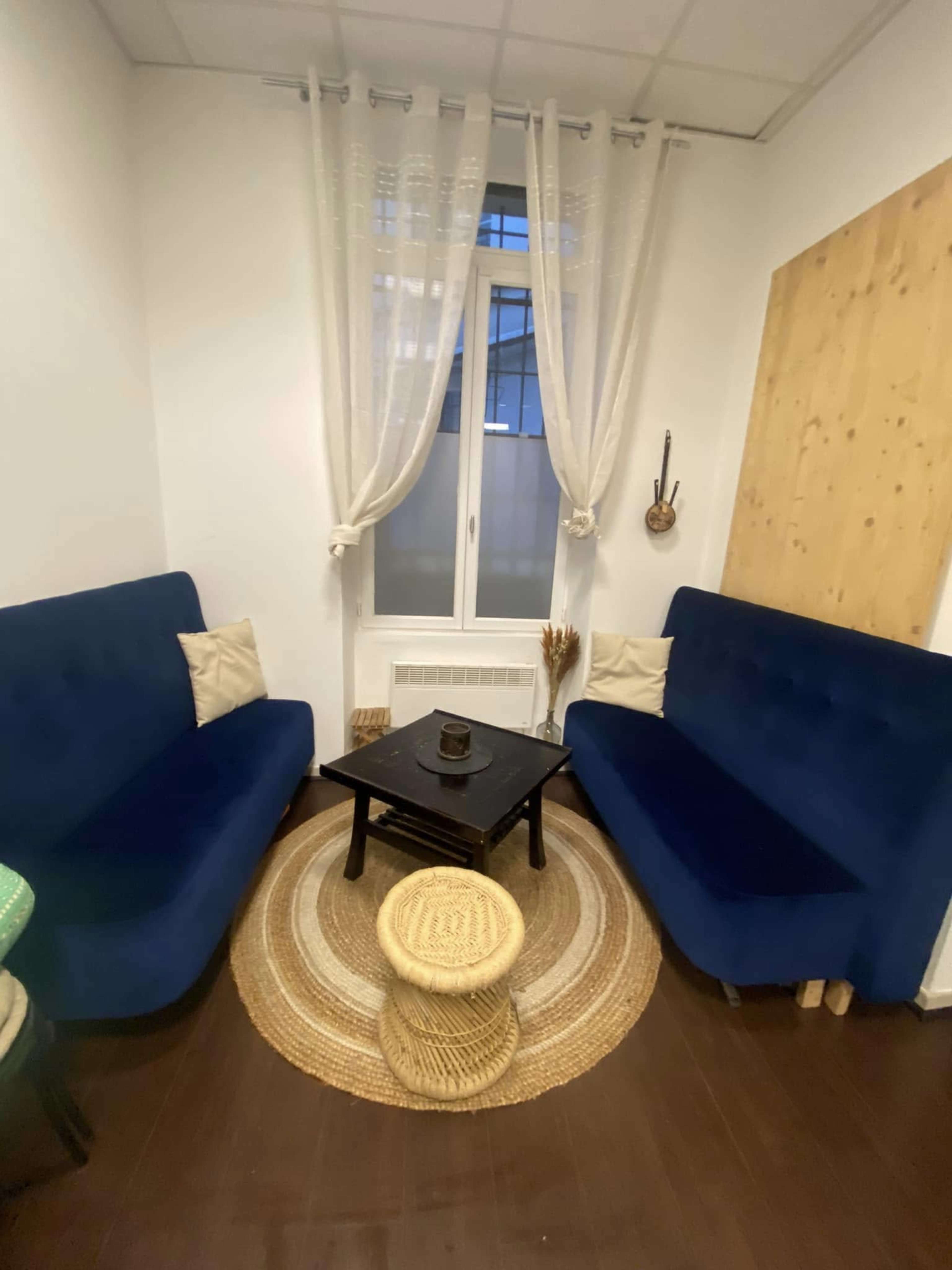 The image shows a small, cozy corner of a room furnished with two blue sofas facing each other around a black wooden table, with a round woven rug underneath and a window with sheer curtains in the background.