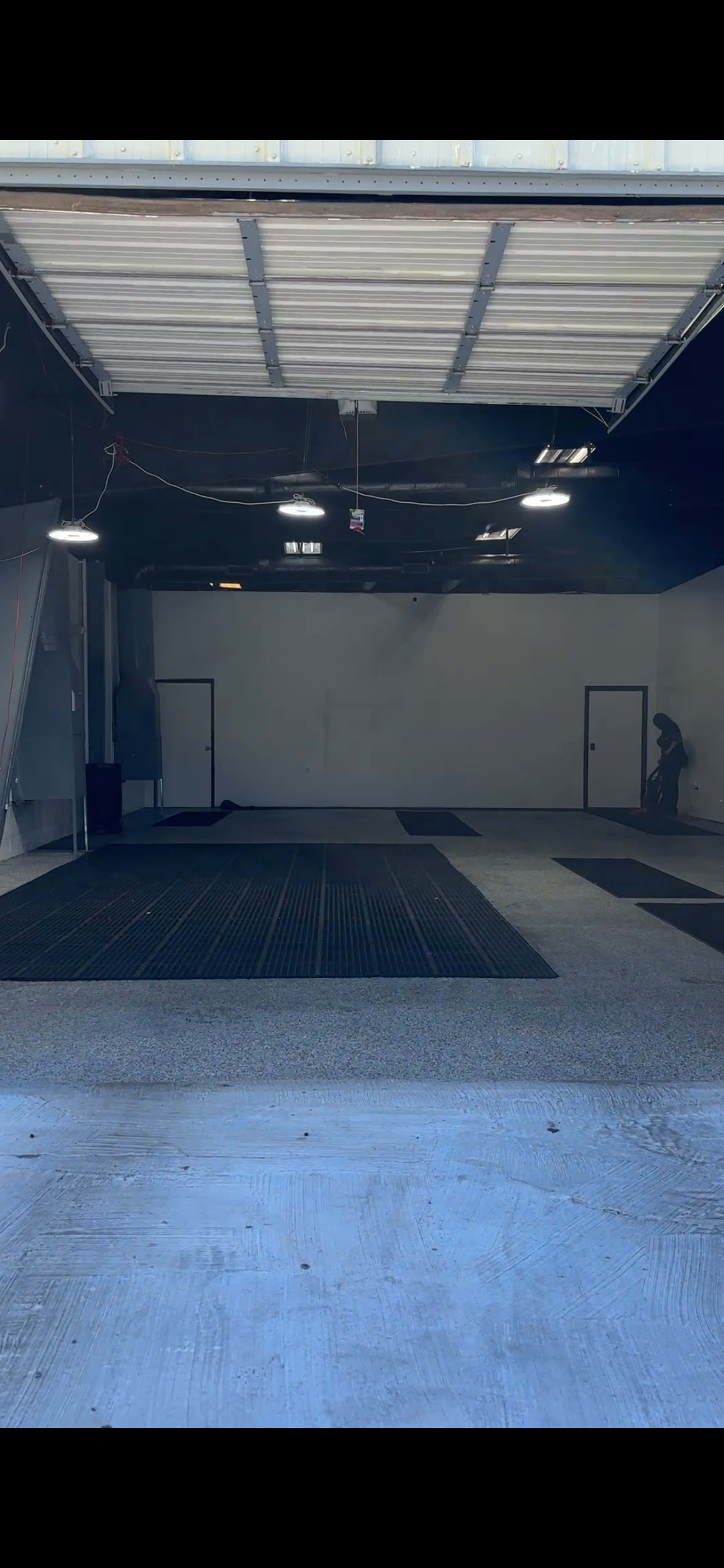 An empty warehouse space with dark rubber mats on the floor and a person sweeping near the back wall.