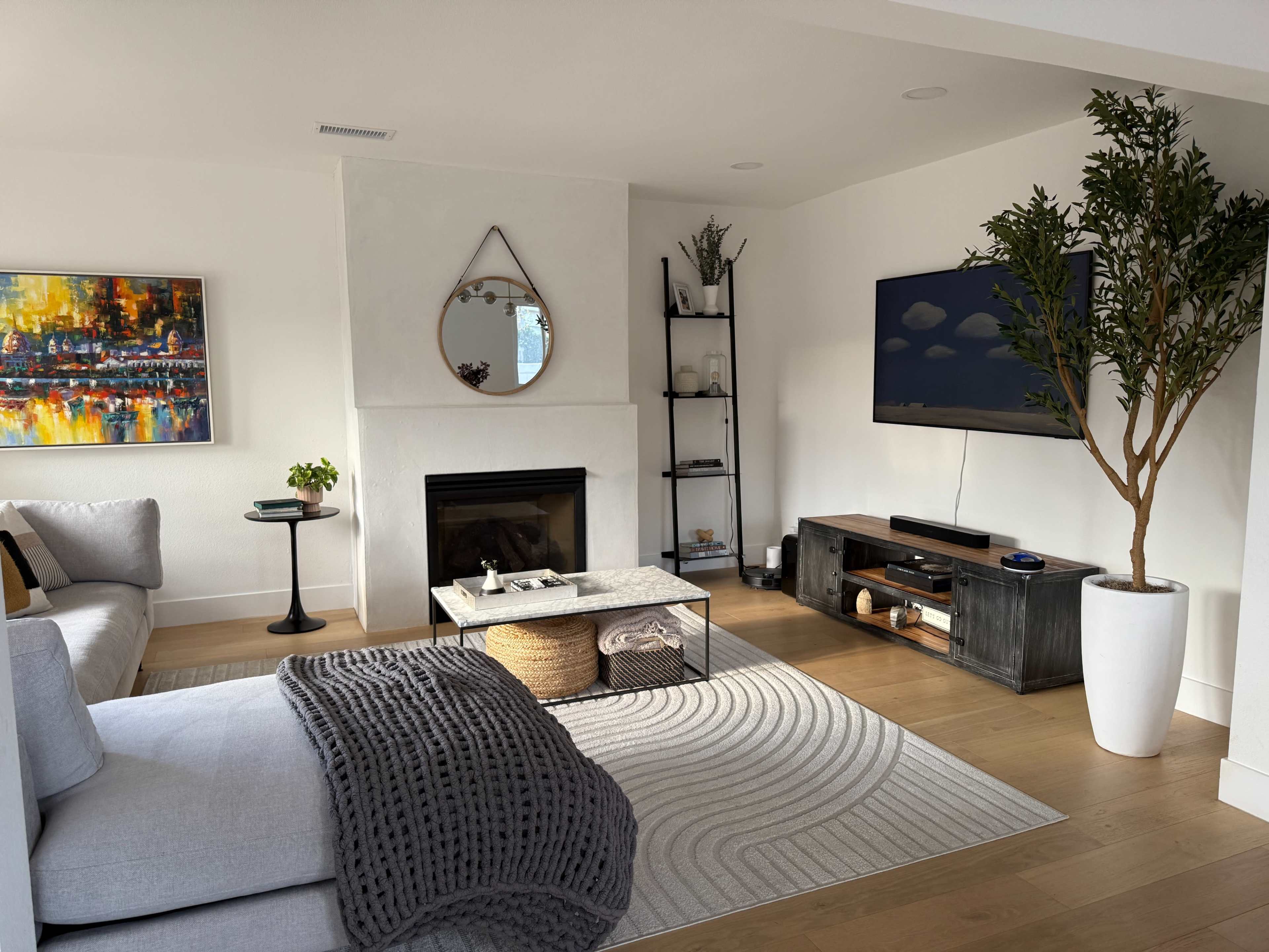 A modern living room features a gray sofa, a round coffee table with woven storage, and a wall-mounted TV, complemented by a small plant and decorative artworks.
