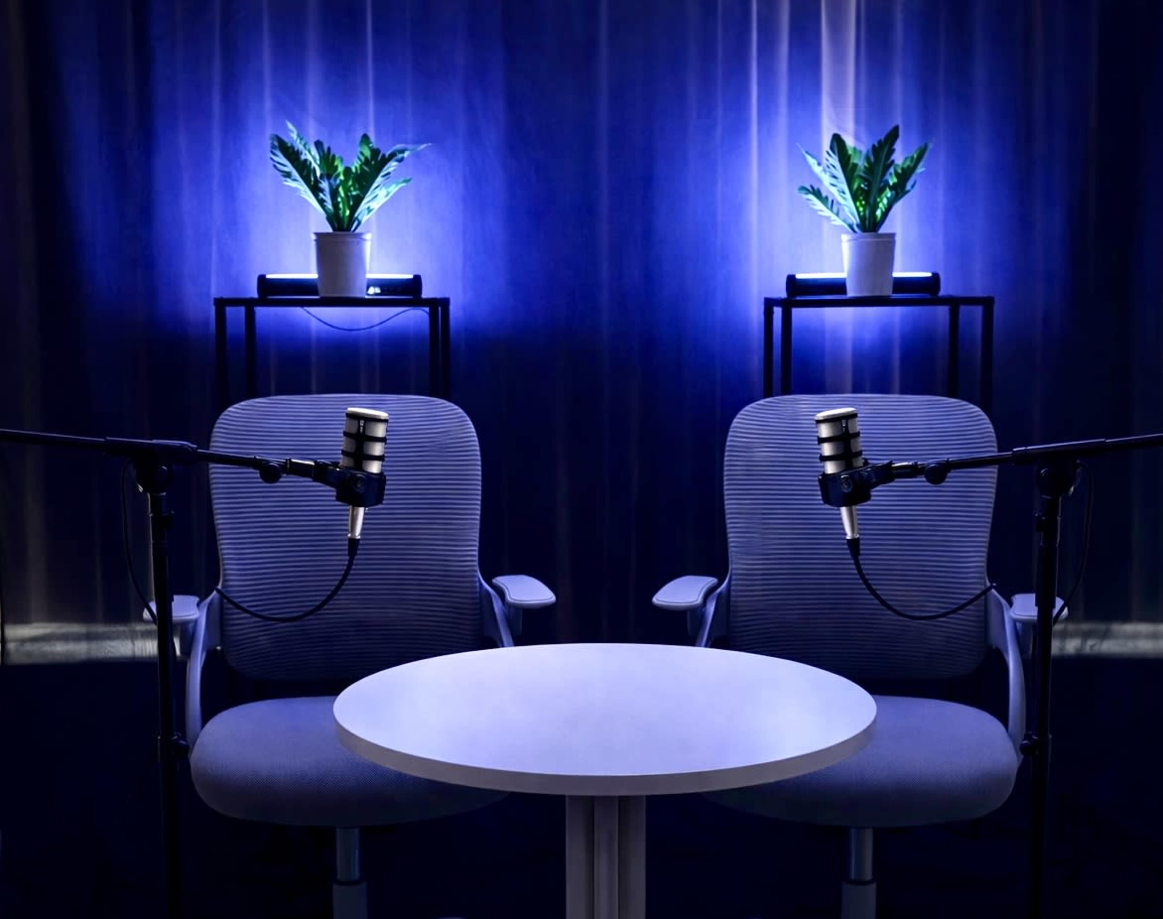 The image shows two chairs with microphones positioned around a small white table, set against a backdrop of blue lighting and potted plants.
