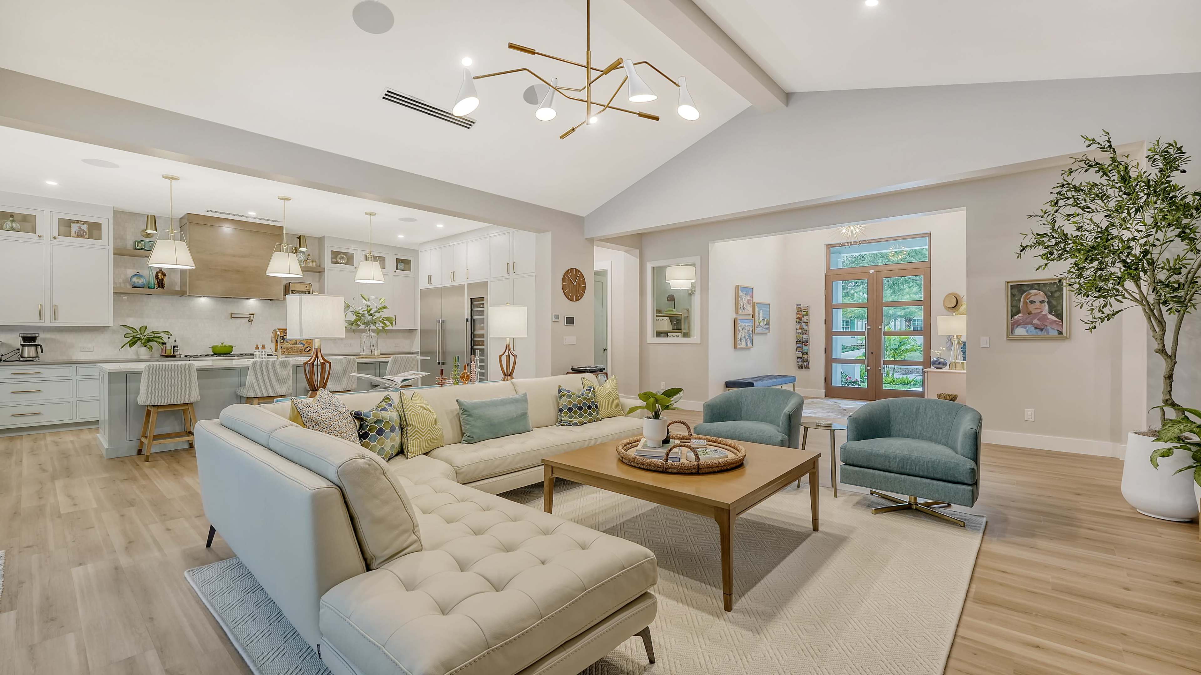 A modern living room features a light-colored sectional sofa, a wooden coffee table, and teal accent chairs, with an open kitchen and large windows in the background.