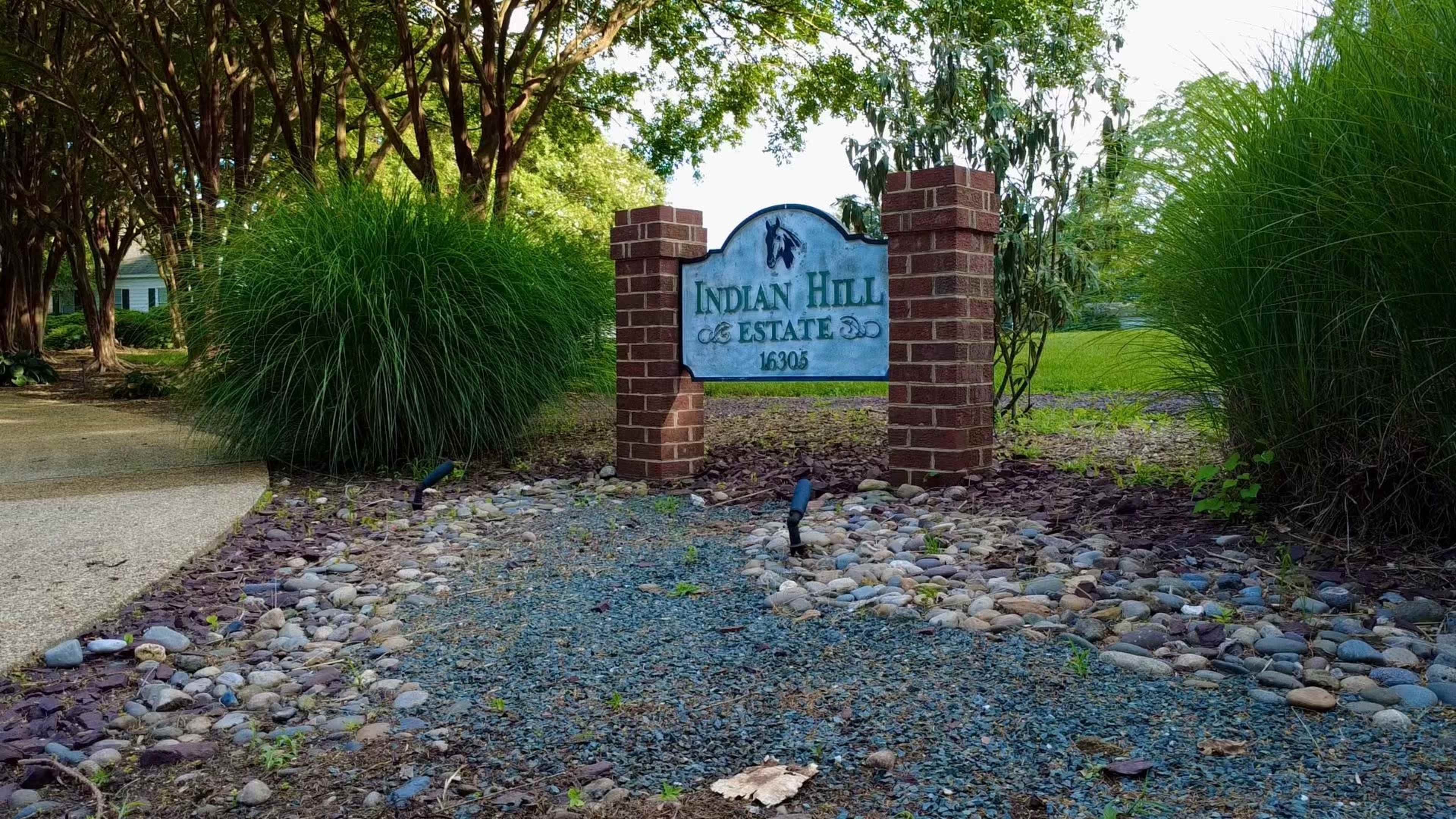 The image shows a stone sign that reads "Indian Hill Estate" surrounded by landscaped greenery, with two birds in the foreground.