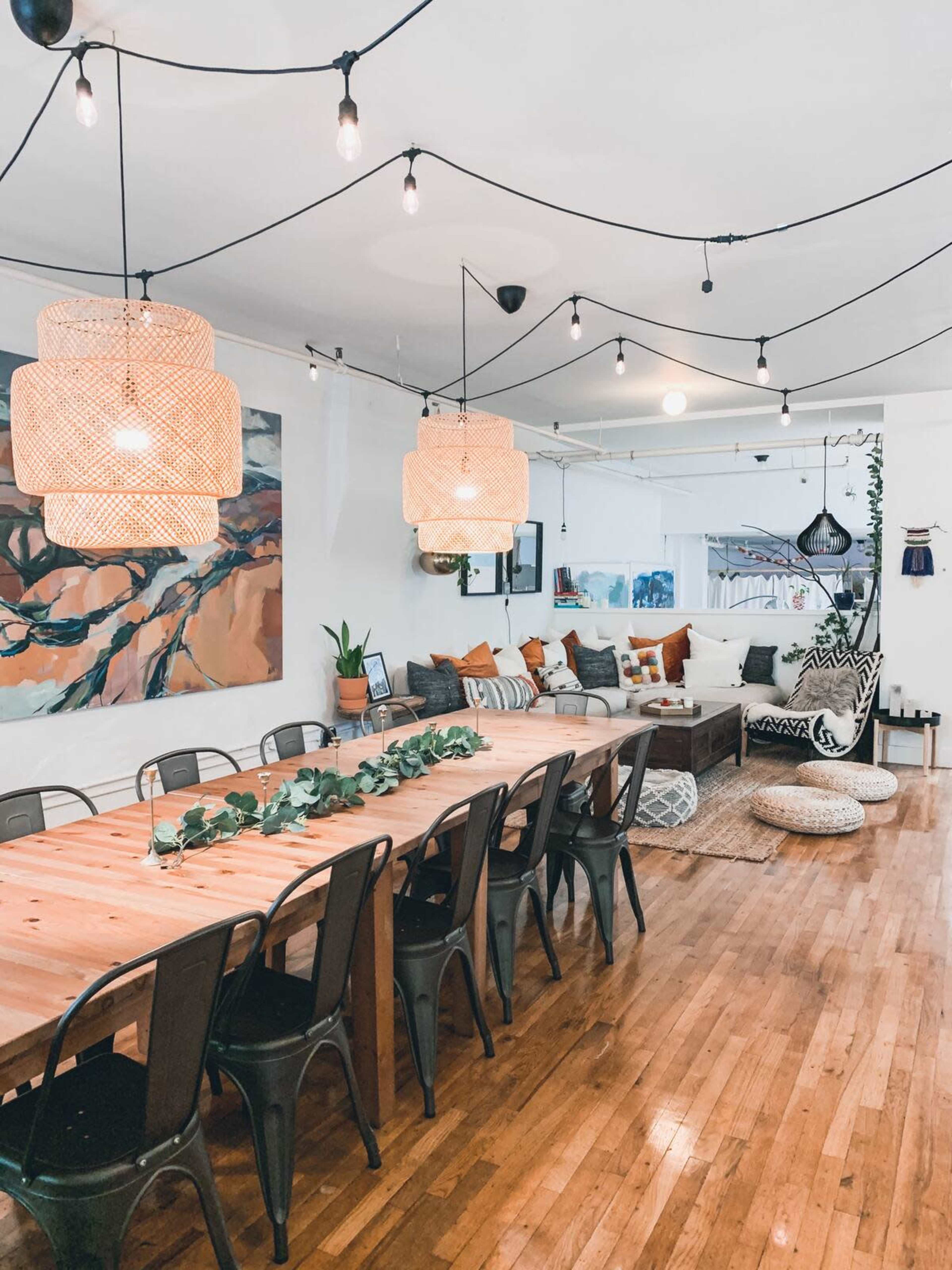 A spacious dining area with a long wooden table surrounded by metal chairs, illuminated by pendant lights and decorated with plants and artwork.