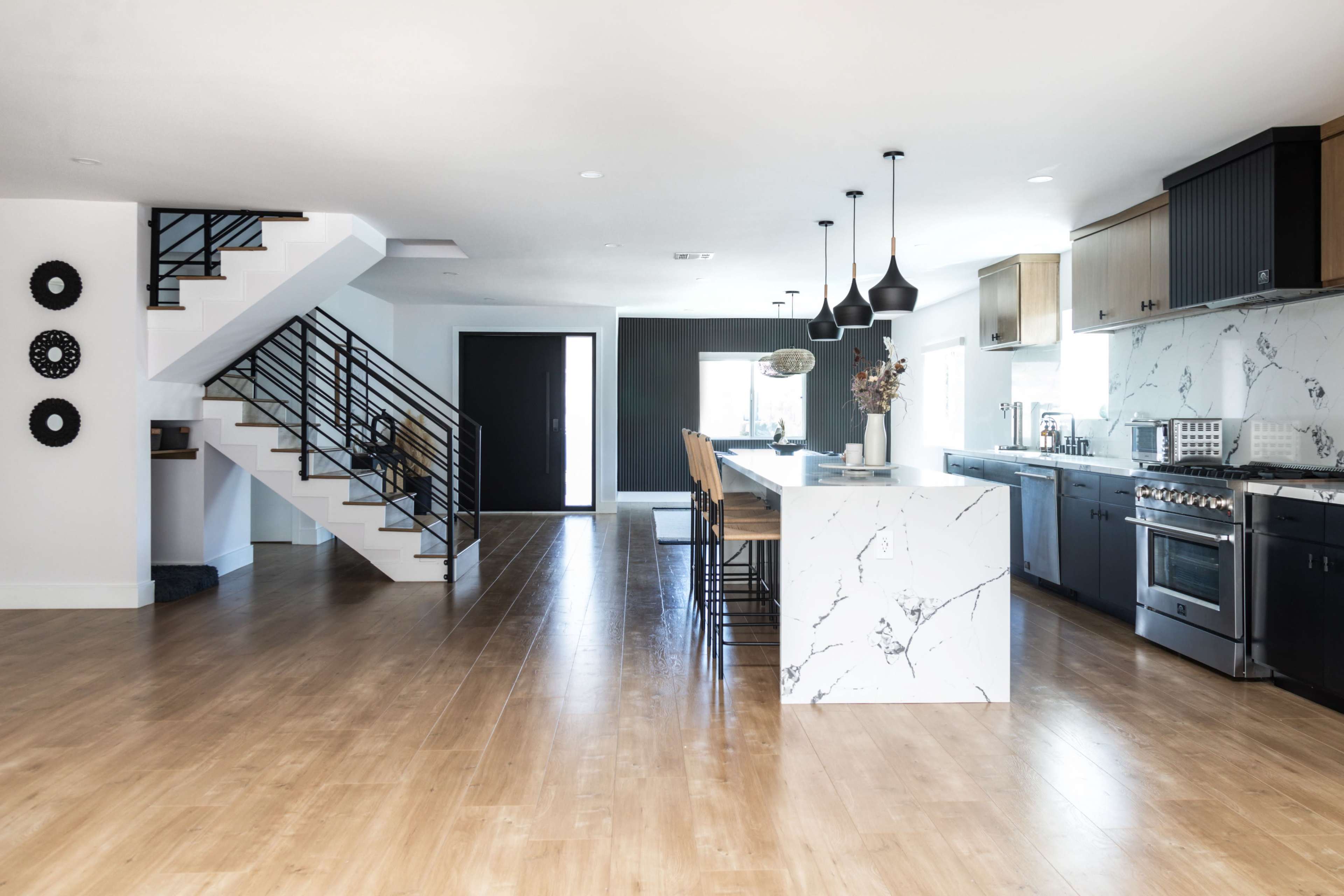 A modern kitchen with a large marble island, adjacent to a staircase and a spacious living area with wooden flooring.