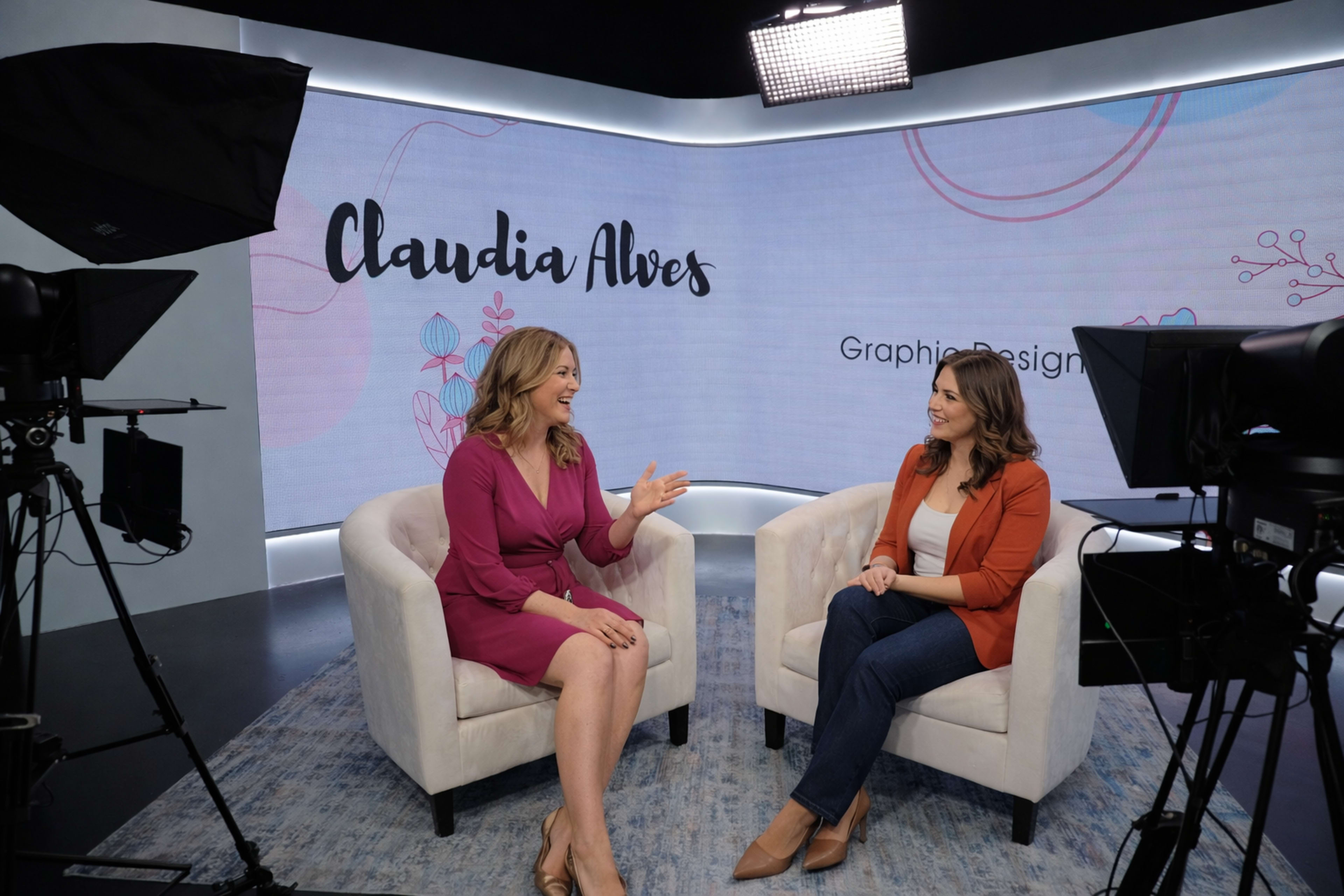 Two women are seated in armchairs facing each other in a studio setting with a backdrop featuring the name "Claudia Alves" and floral graphics.