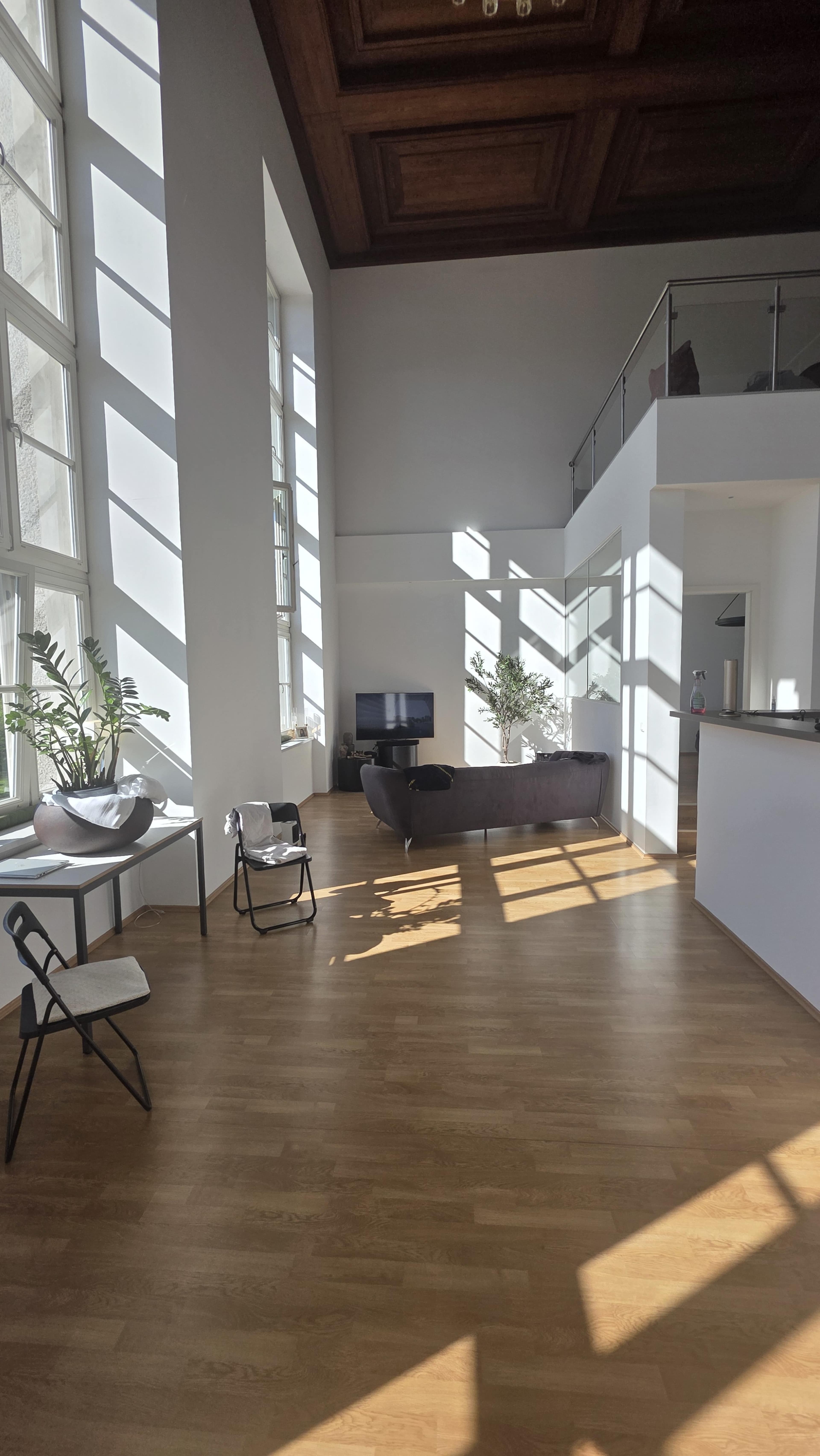 The image shows a spacious, modern living area with large windows casting diagonal shadows on the wooden floor.