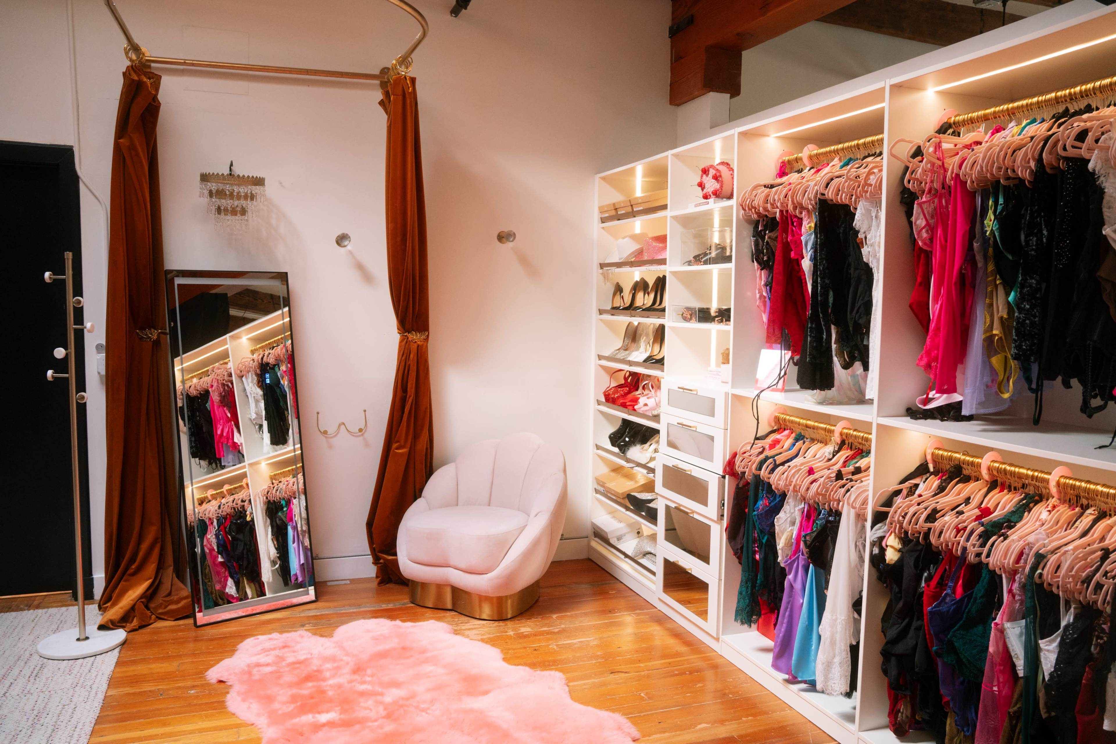The image shows a stylish dressing room featuring an arranged collection of colorful clothing on hangers, a soft pink chair, a mirror, and a plush rug.