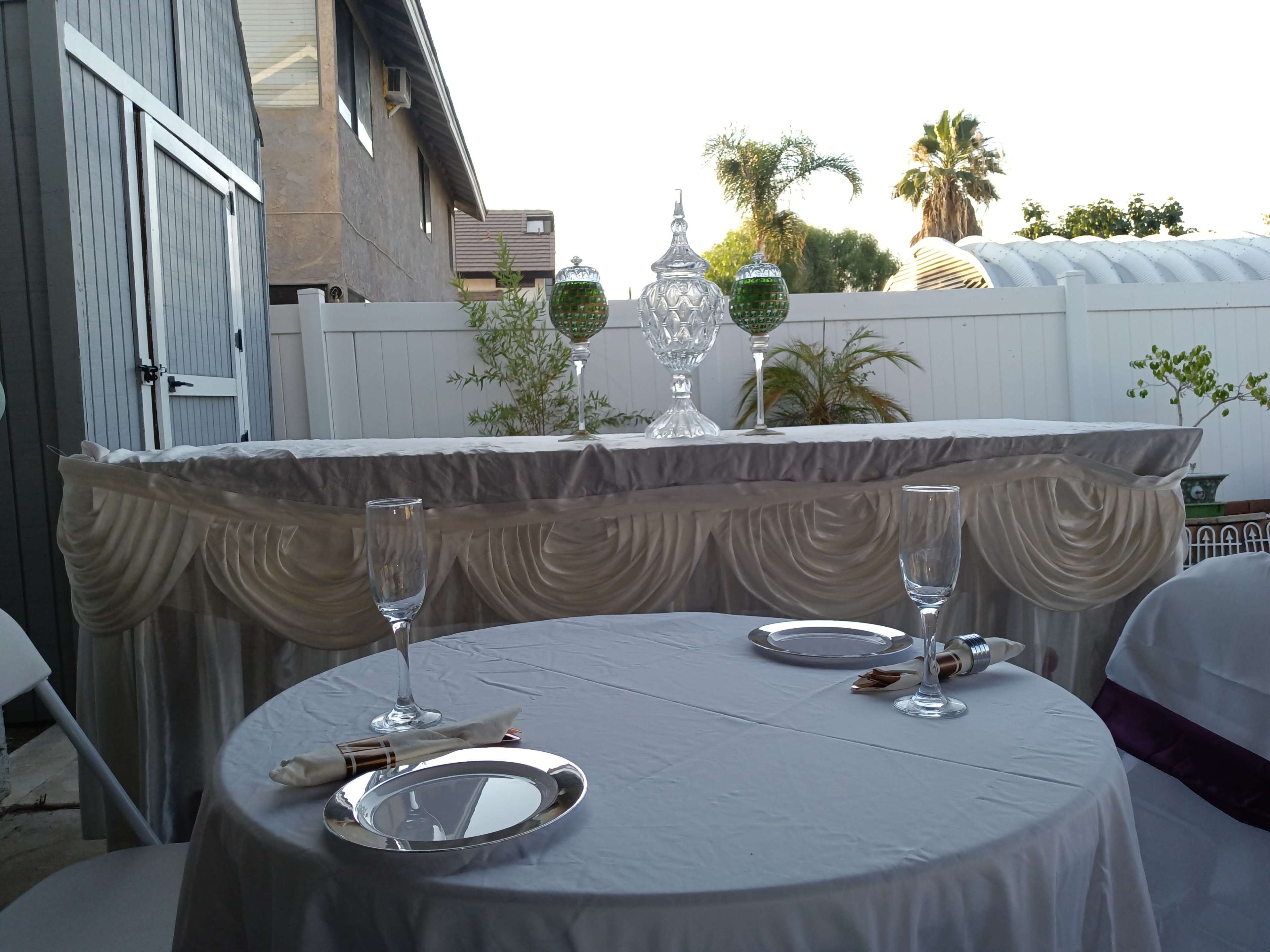 A decorated outdoor table setting features a draped tablecloth, crystal centerpiece, and glassware, surrounded by plants and a white fence.