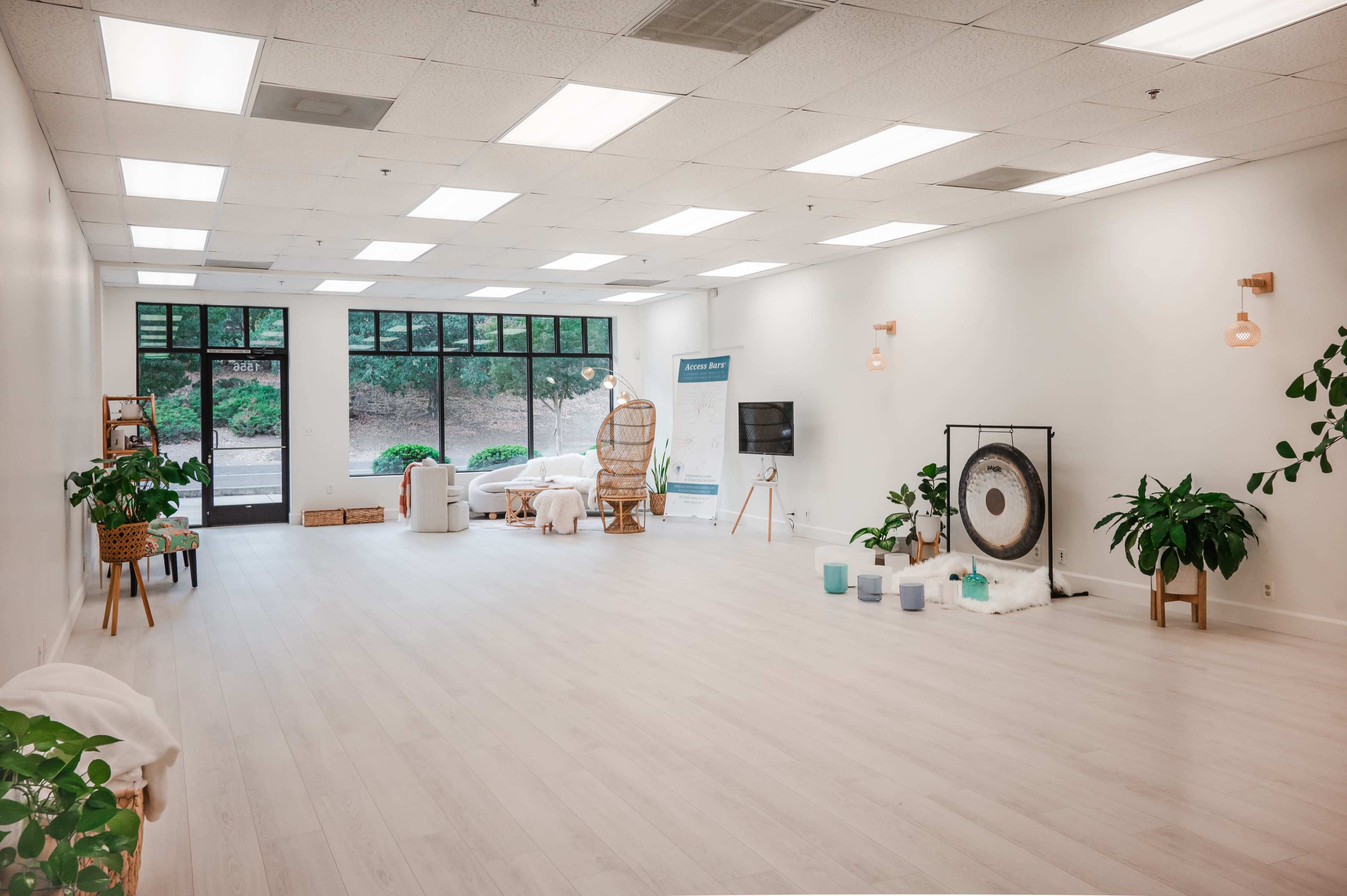 An airy, minimalist wellness studio features a large open space with wooden floors, potted plants, a gong, and natural lighting from large windows.