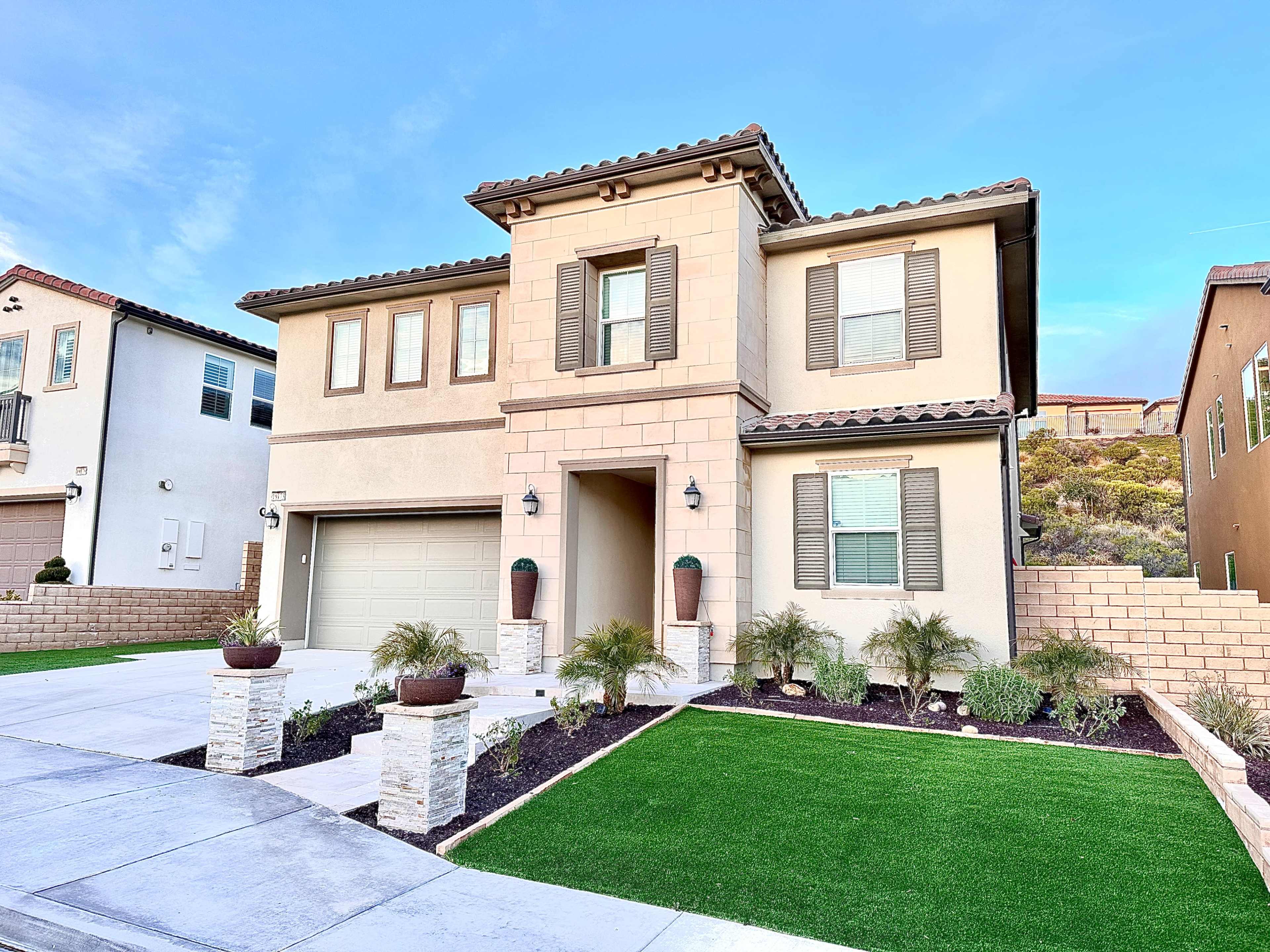 A two-story house with a stone facade, a two-car garage, and well-maintained landscaping featuring potted plants and artificial grass.