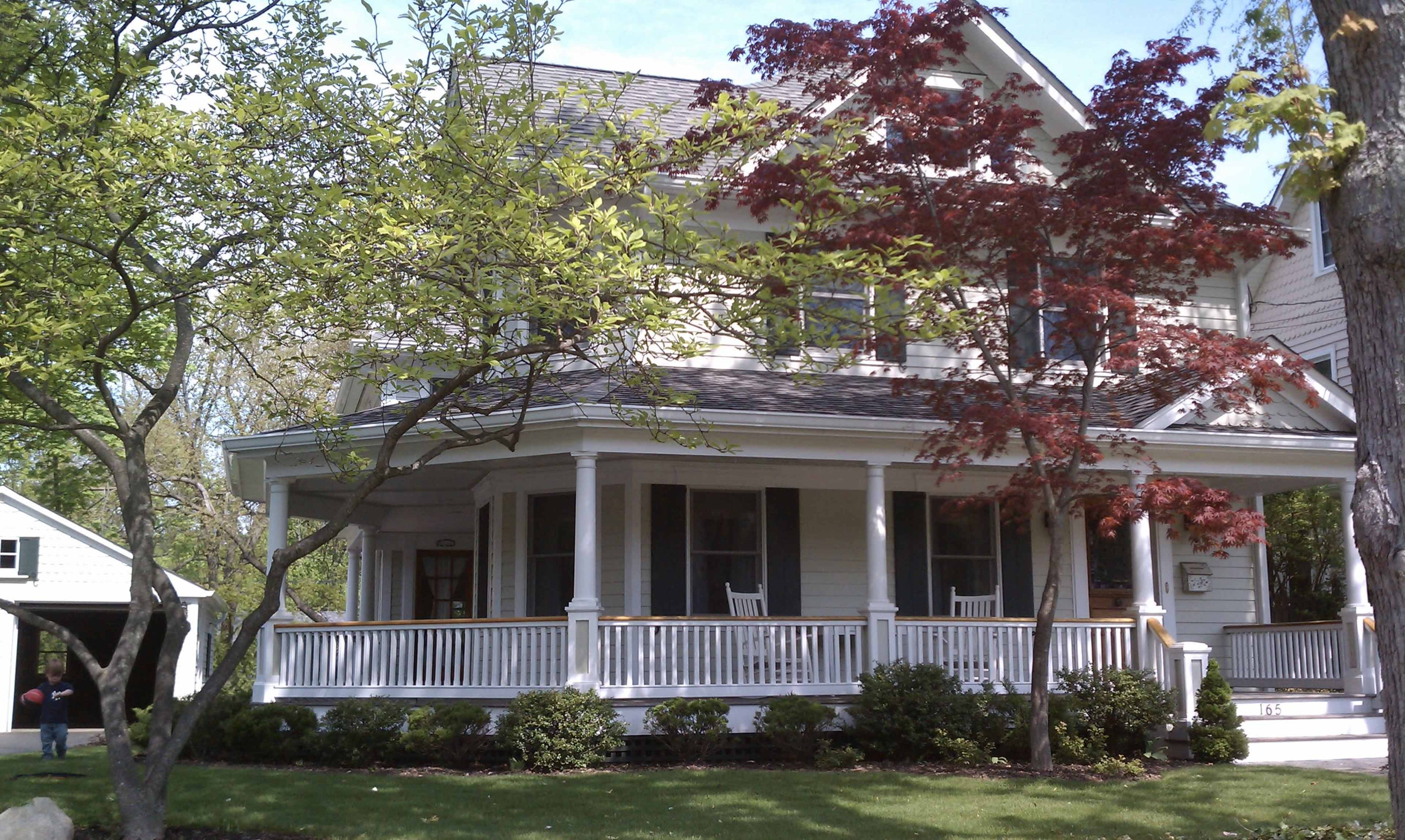 A two-story house with a wraparound porch and landscaped gardens featuring a small red tree and green foliage.