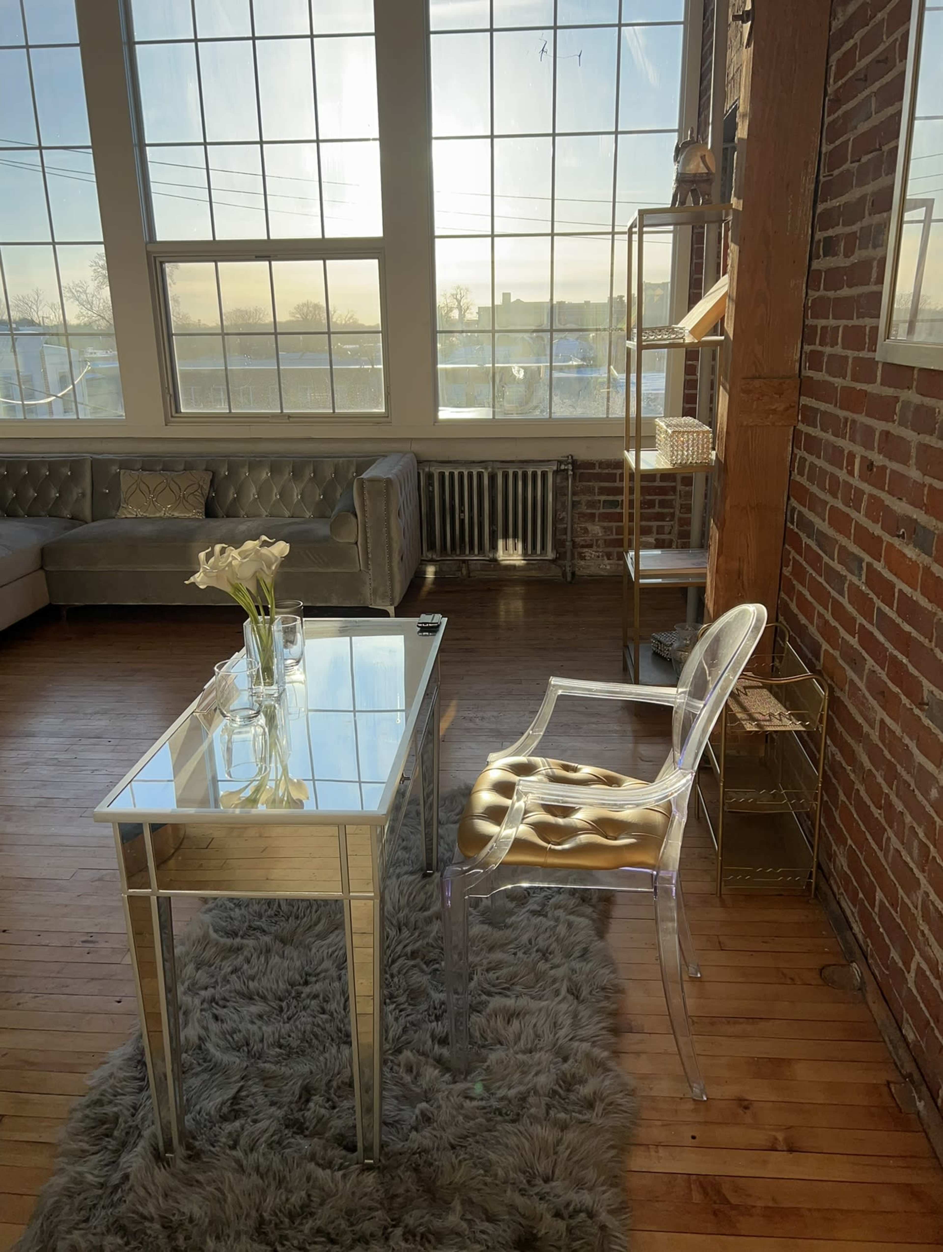 A glass-top table and a clear acrylic chair are set beside a window in a room with exposed brick walls and wooden floors, featuring a cozy rug and a sofa in the background.