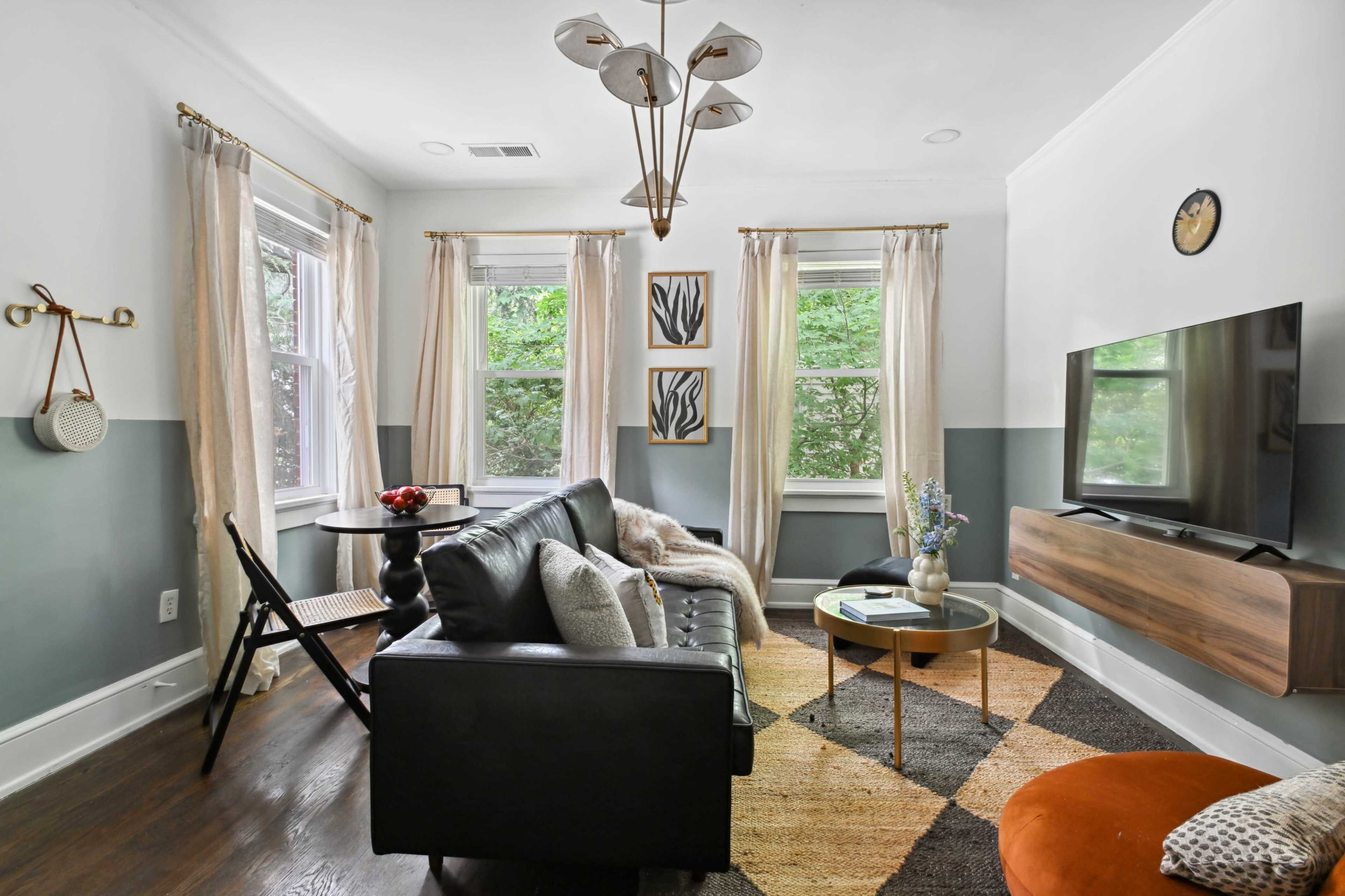 The living room features a black leather couch, a circular coffee table, and a flat-screen TV mounted on the wall, with natural light streaming through two windows.