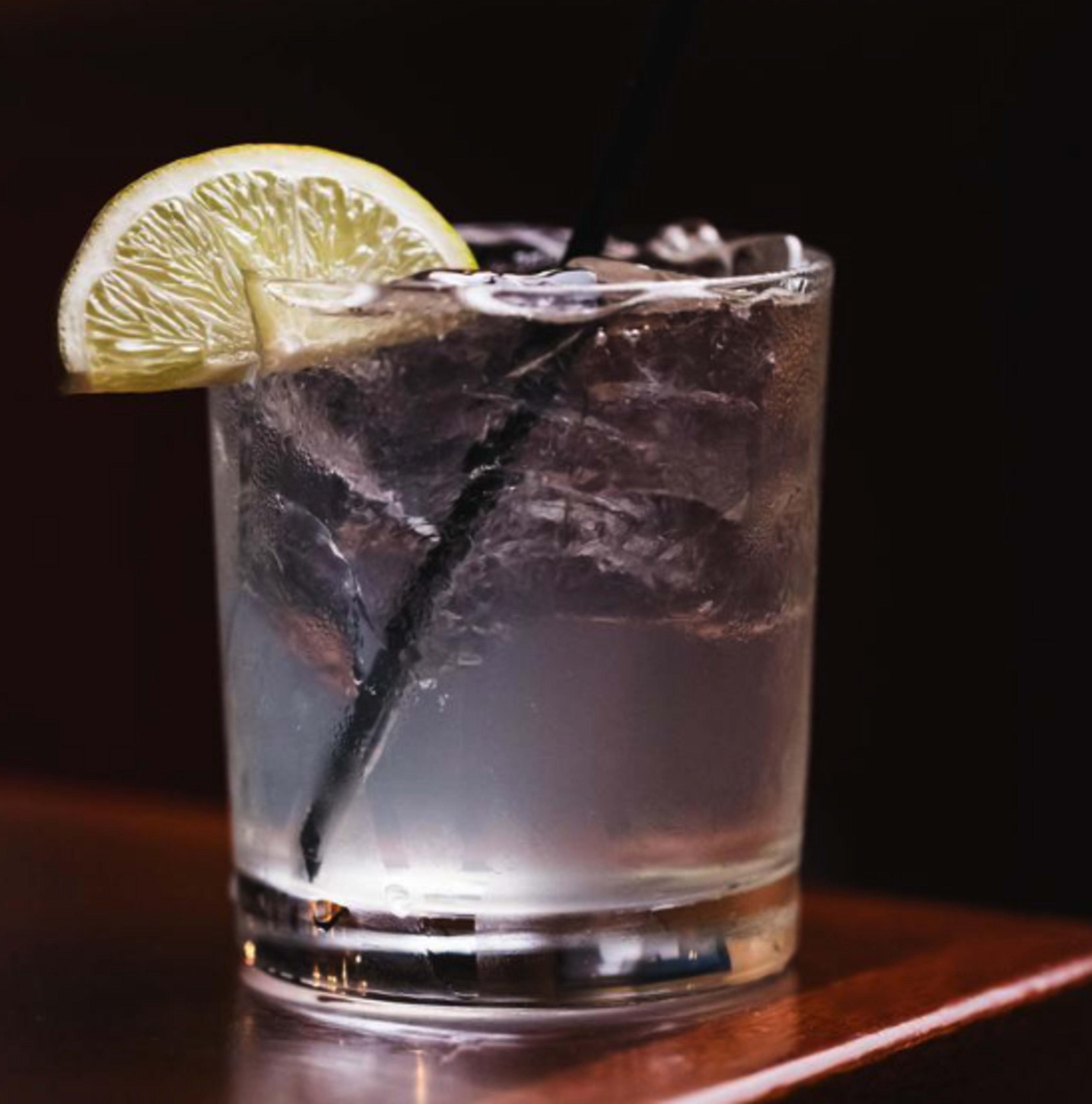 A glass of clear liquid with ice and a slice of lemon is placed on a wooden surface.