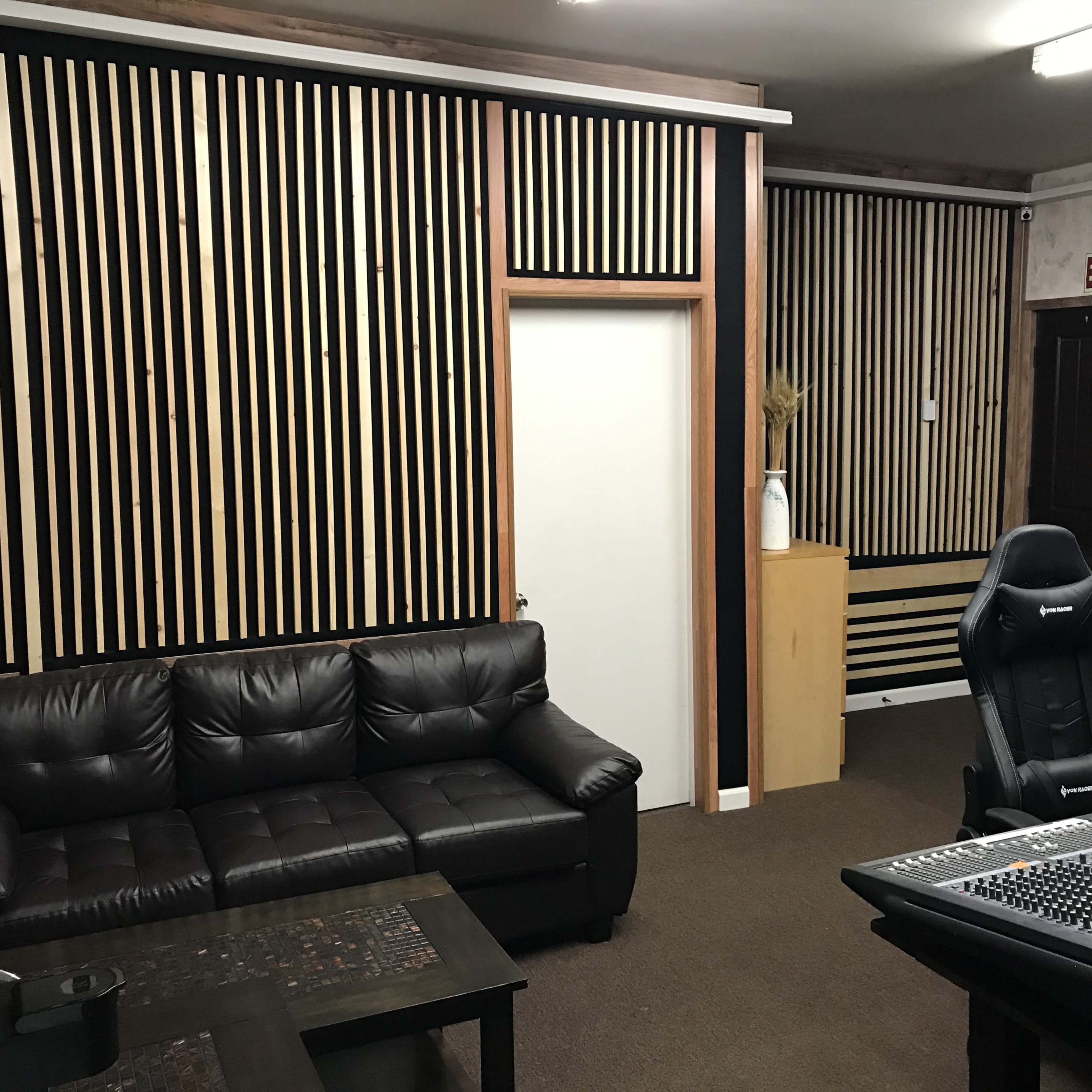 The image shows a room with a black and wood slatted wall design, featuring a dark leather sofa, a wooden cabinet, and a desk with audio equipment.