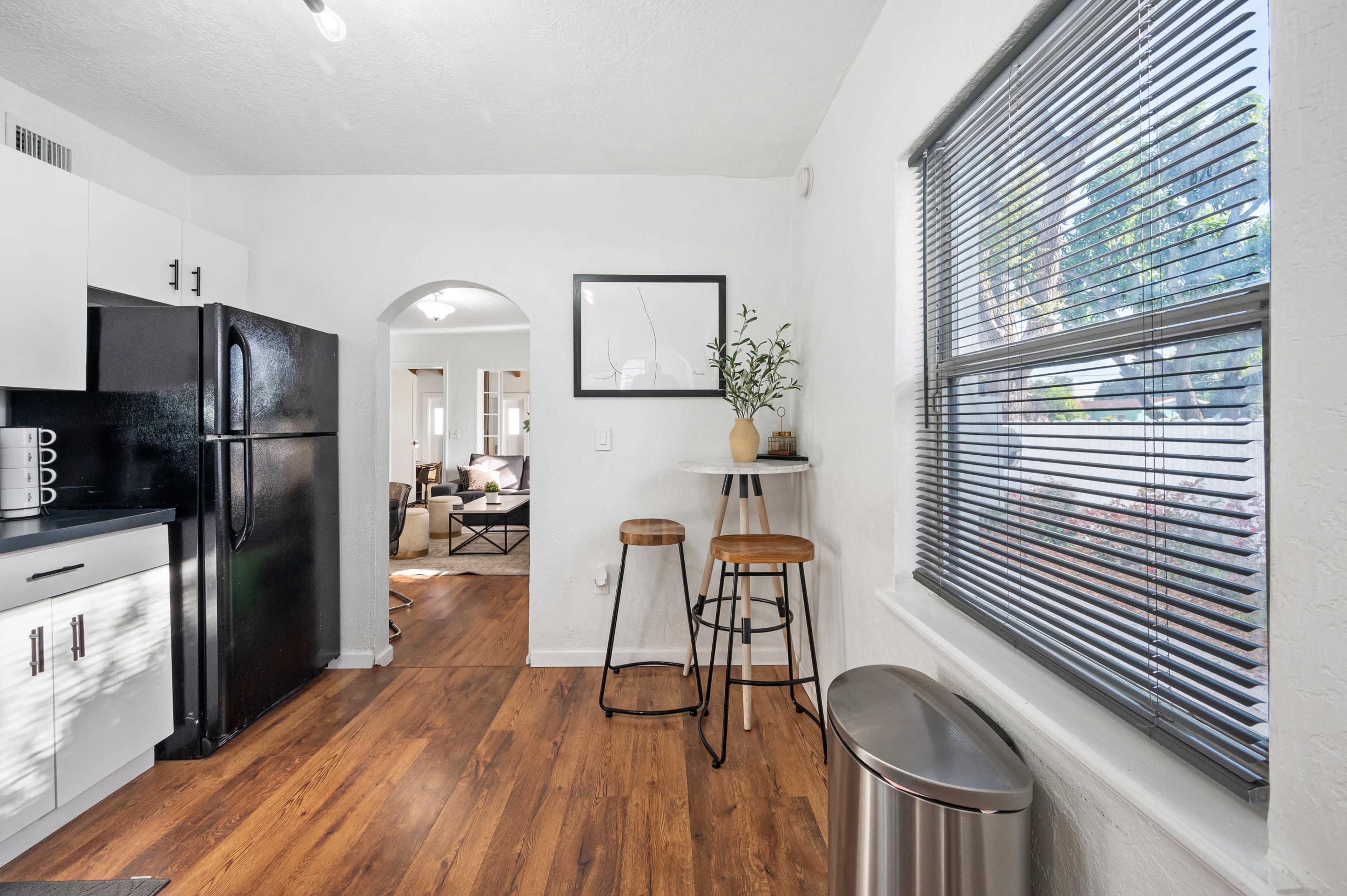 The image shows a kitchen area with black appliances, a small dining setup with two stools, and a view into a living space through an archway.
