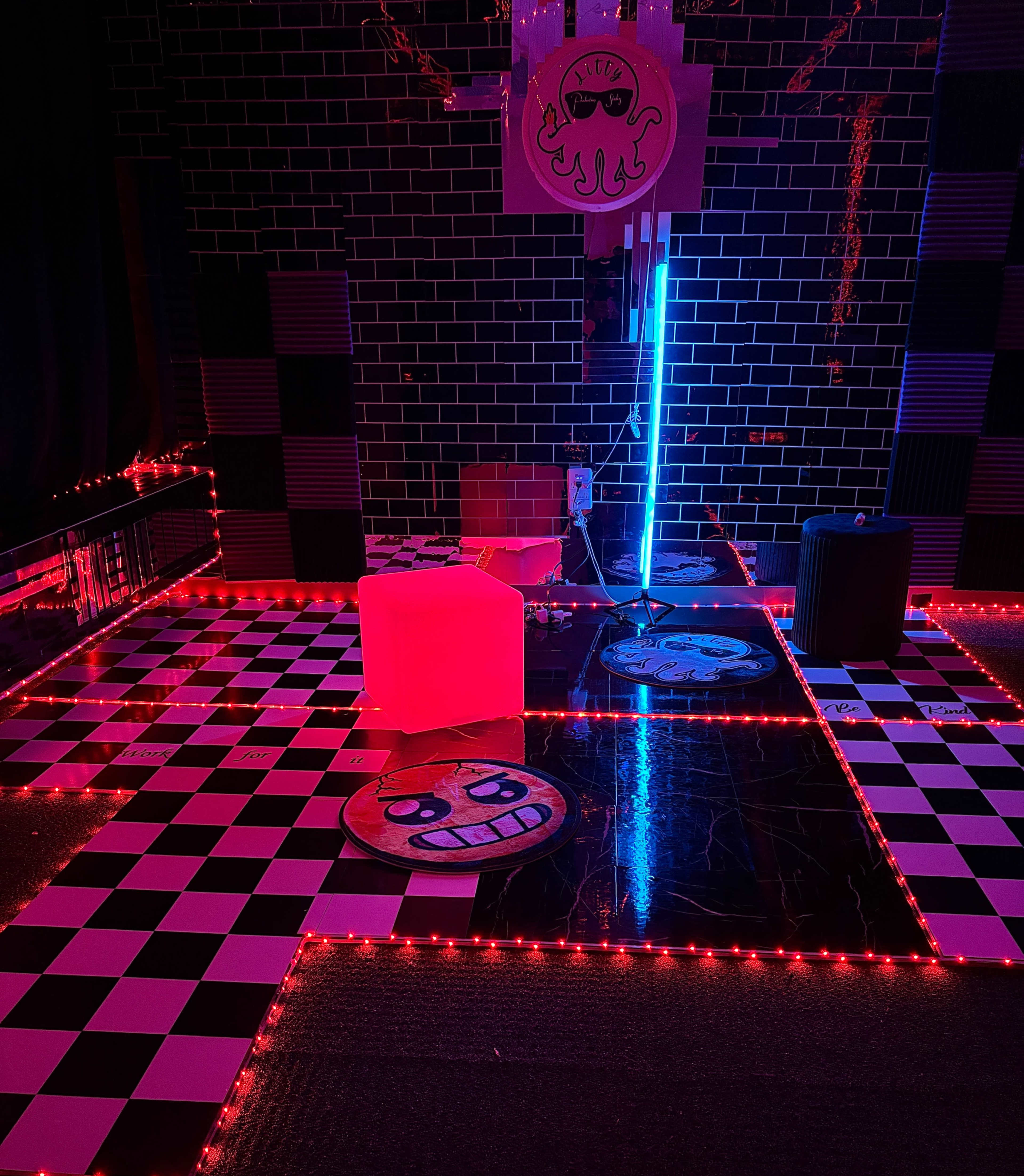 The image depicts a room with a checkered black and white floor, illuminated by red LED lights, featuring a neon blue light and various decorative elements, including round mats and a wall sign.