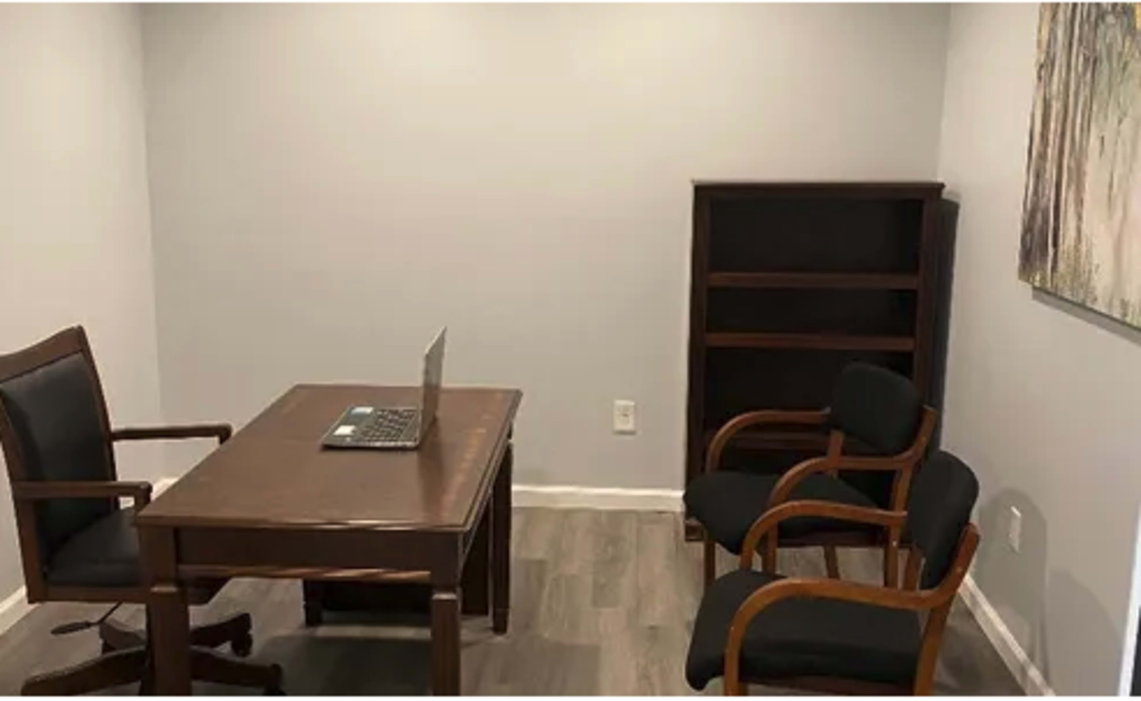 A simple office space featuring a wooden desk with a laptop, two chairs, and a bookshelf against a gray wall.