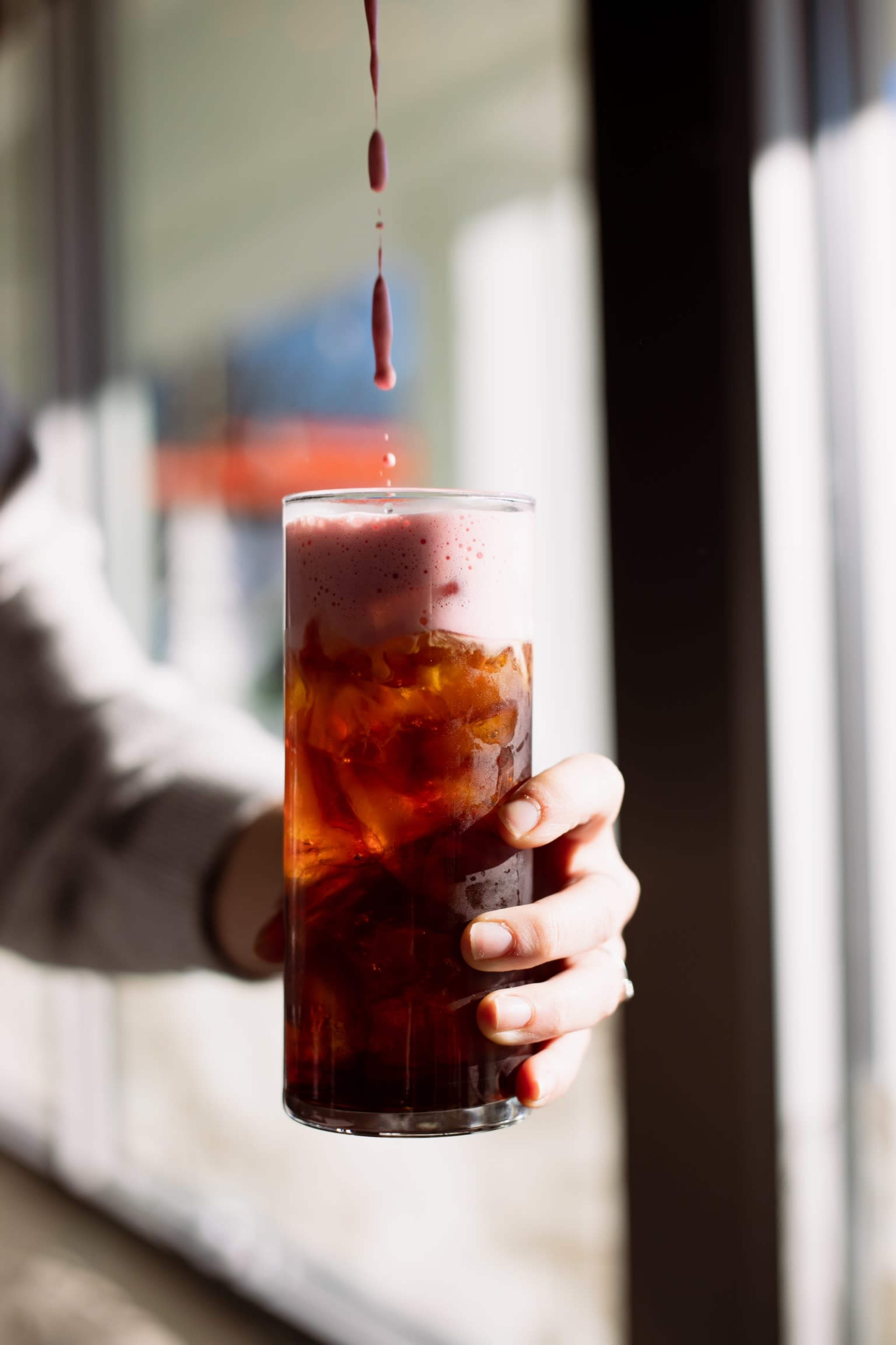 A hand holds a tall glass filled with iced tea or coffee as a drop of syrup drips into it.