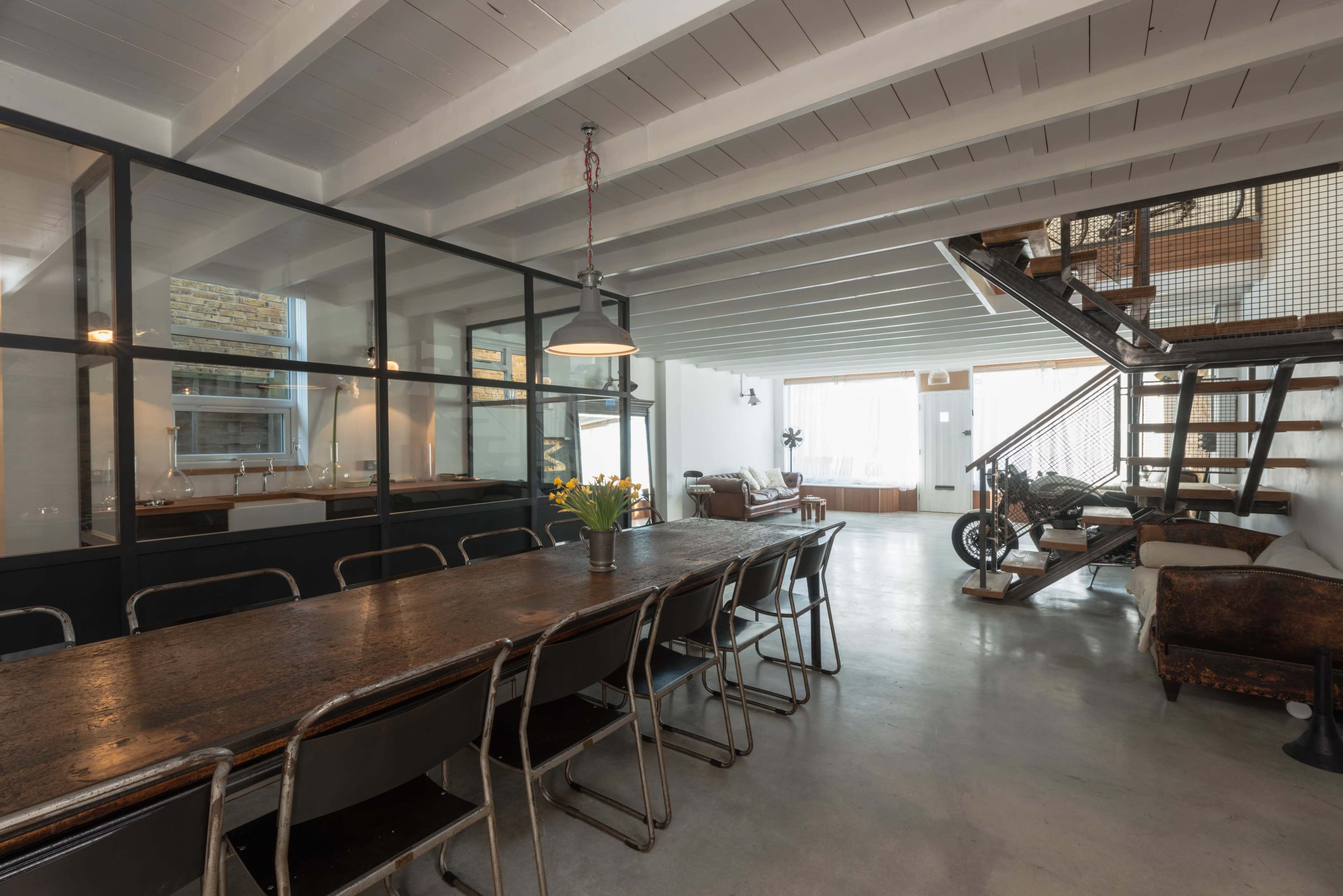 A modern, open-plan living space featuring a long dining table, glass partition, a staircase, and a mix of seating areas.