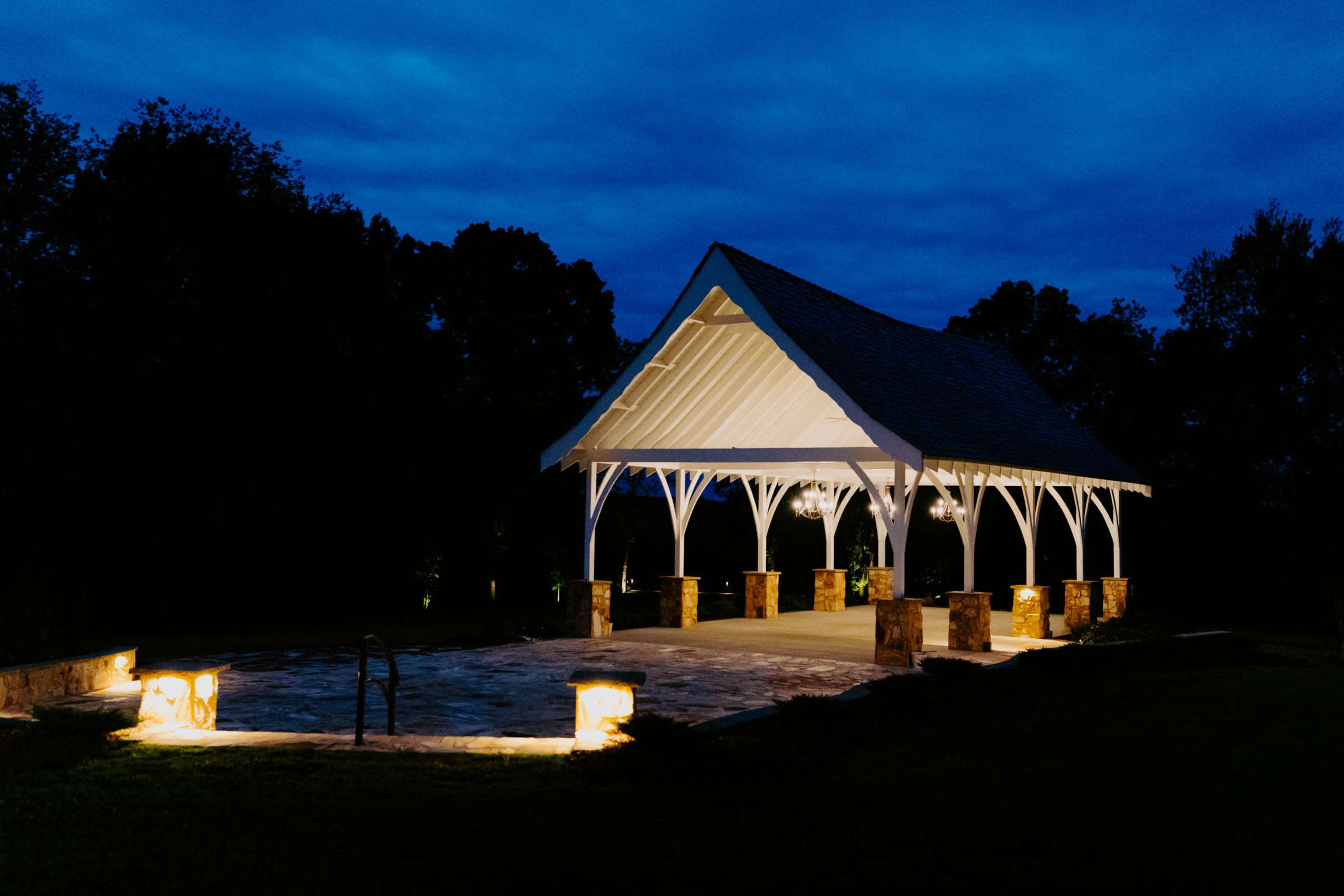 A white wooden pavilion with stone accents is illuminated at night against a cloudy blue sky.
