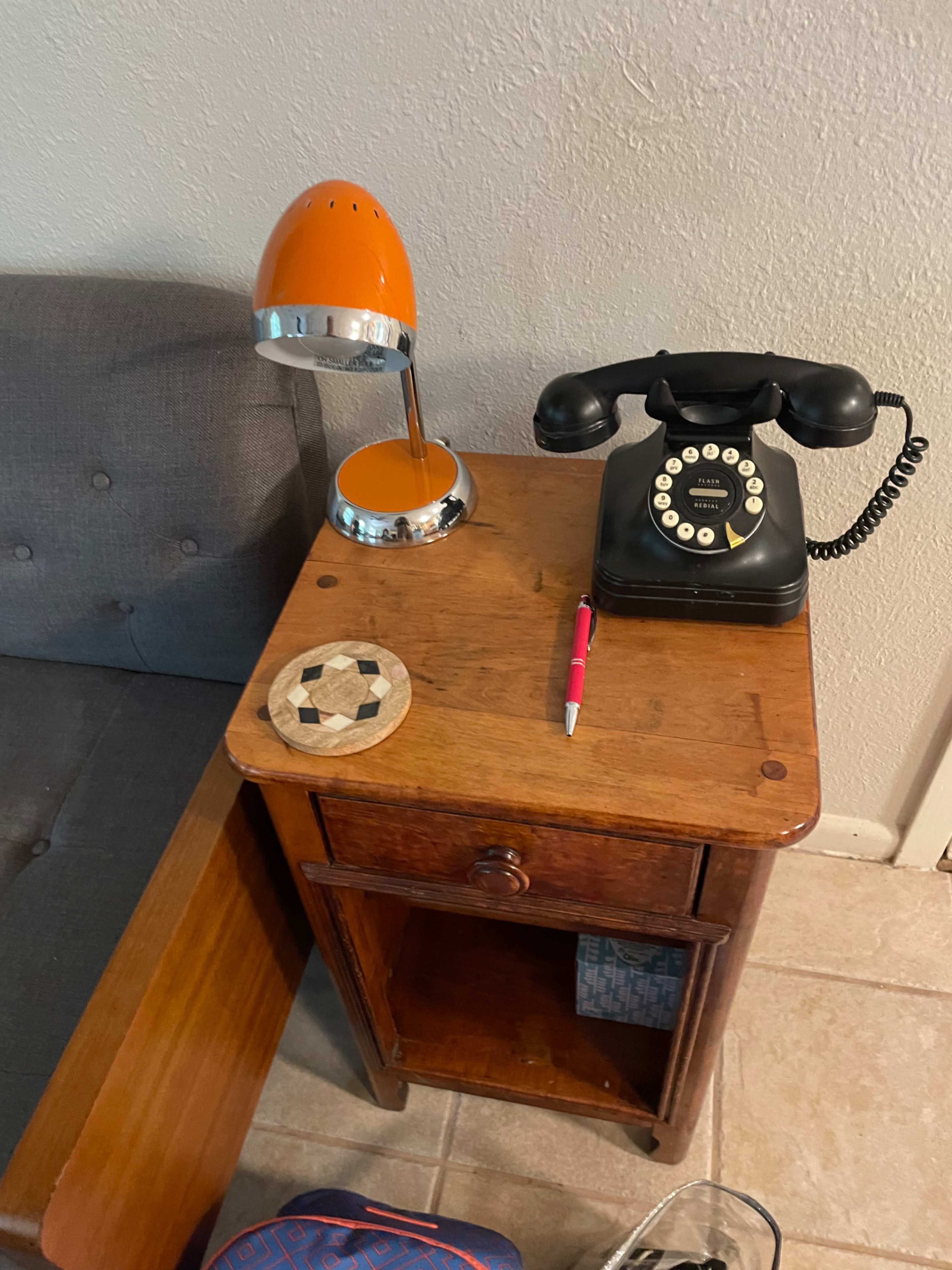A vintage black rotary telephone and a bright orange desk lamp are placed on a wooden side table next to a couch.