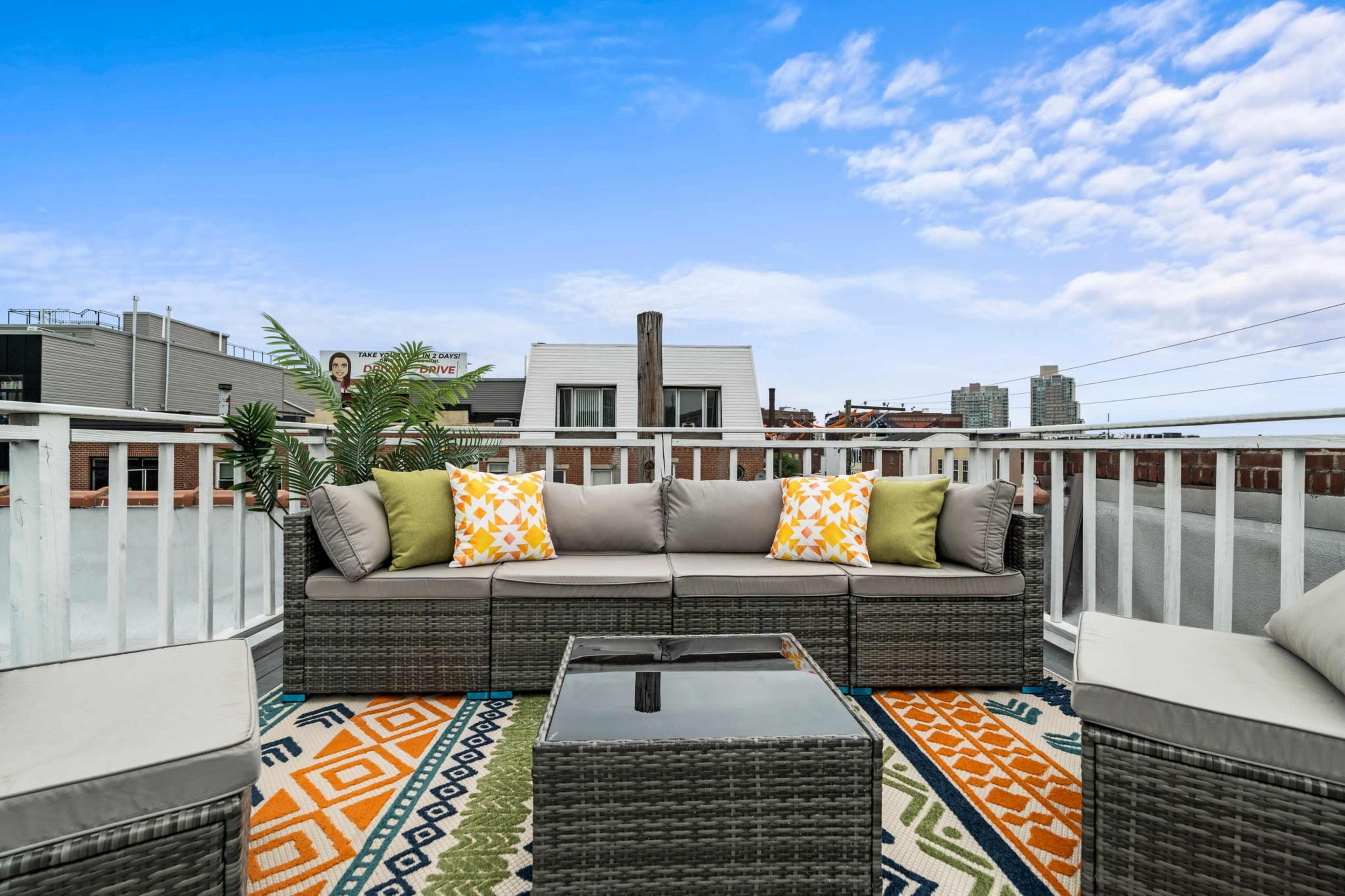 A rooftop terrace features a sectional sofa with colorful cushions, a glass-topped table, and a decorative outdoor rug under a clear blue sky.