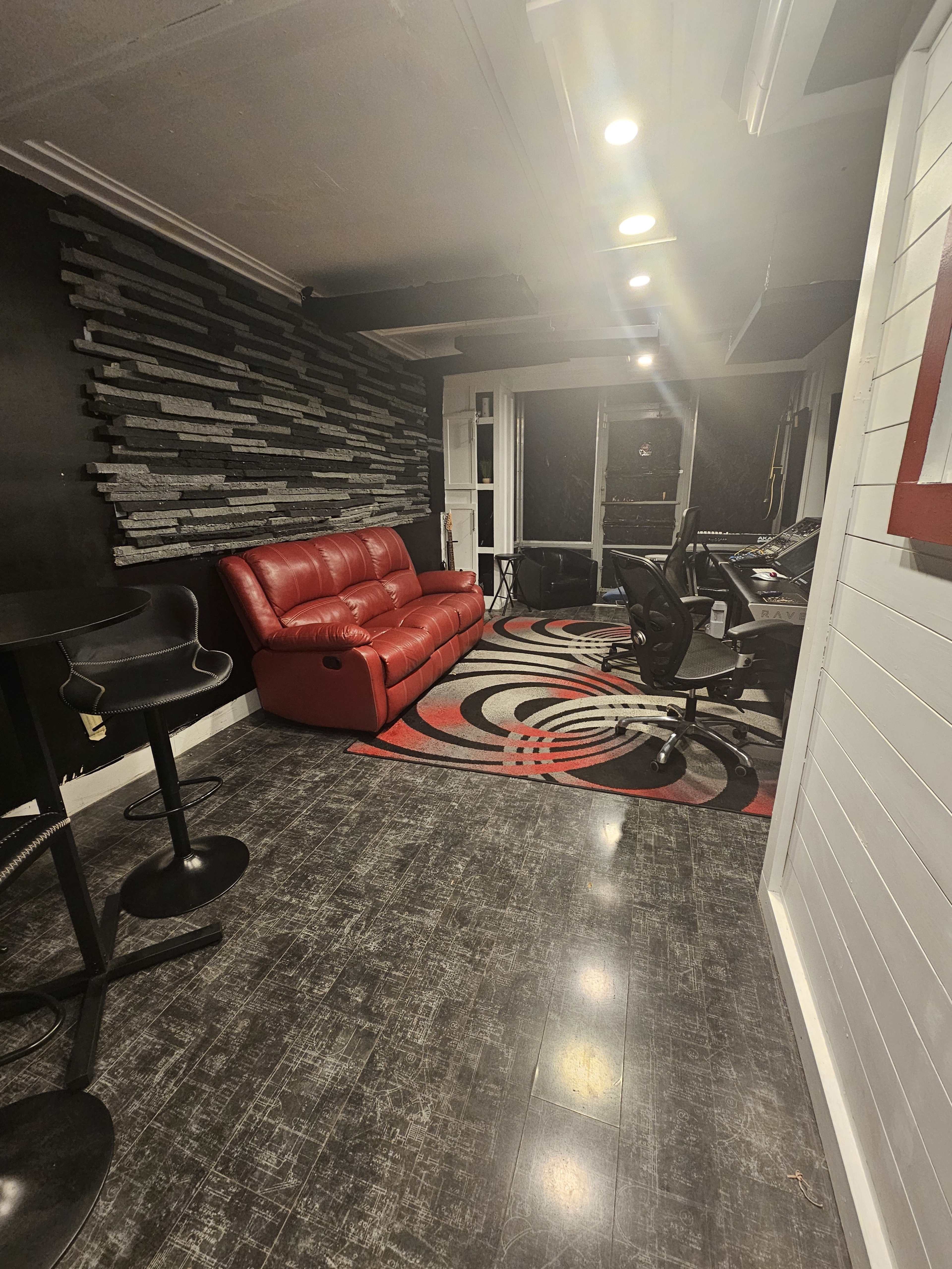 The image shows a modern room with a red leather couch, a black bar stool, and a desk with office chairs, all set against a textured stone wall and a patterned rug.
