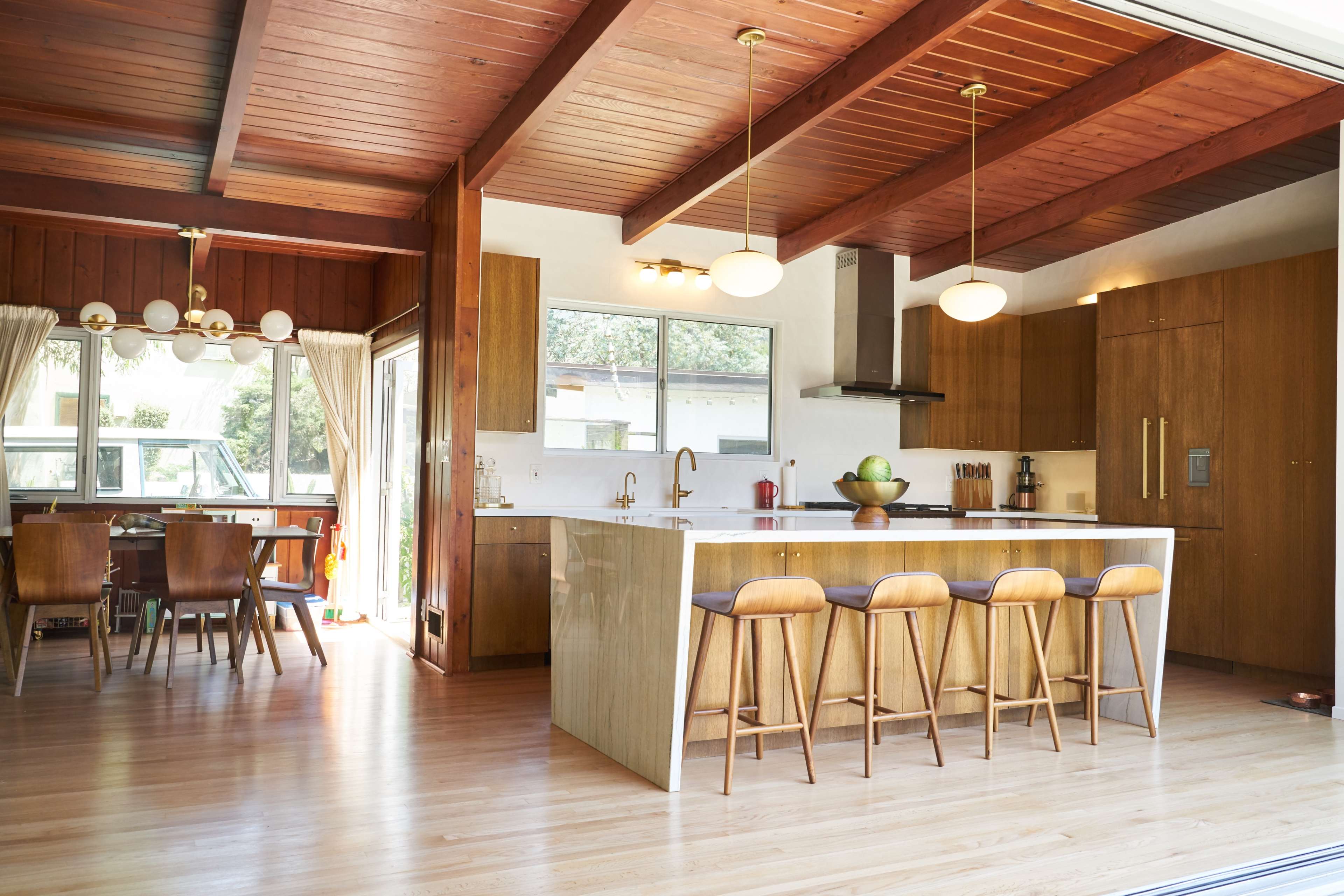The image depicts a modern kitchen with a large island and wooden cabinetry, adjacent to a dining area with a wooden table and chairs.