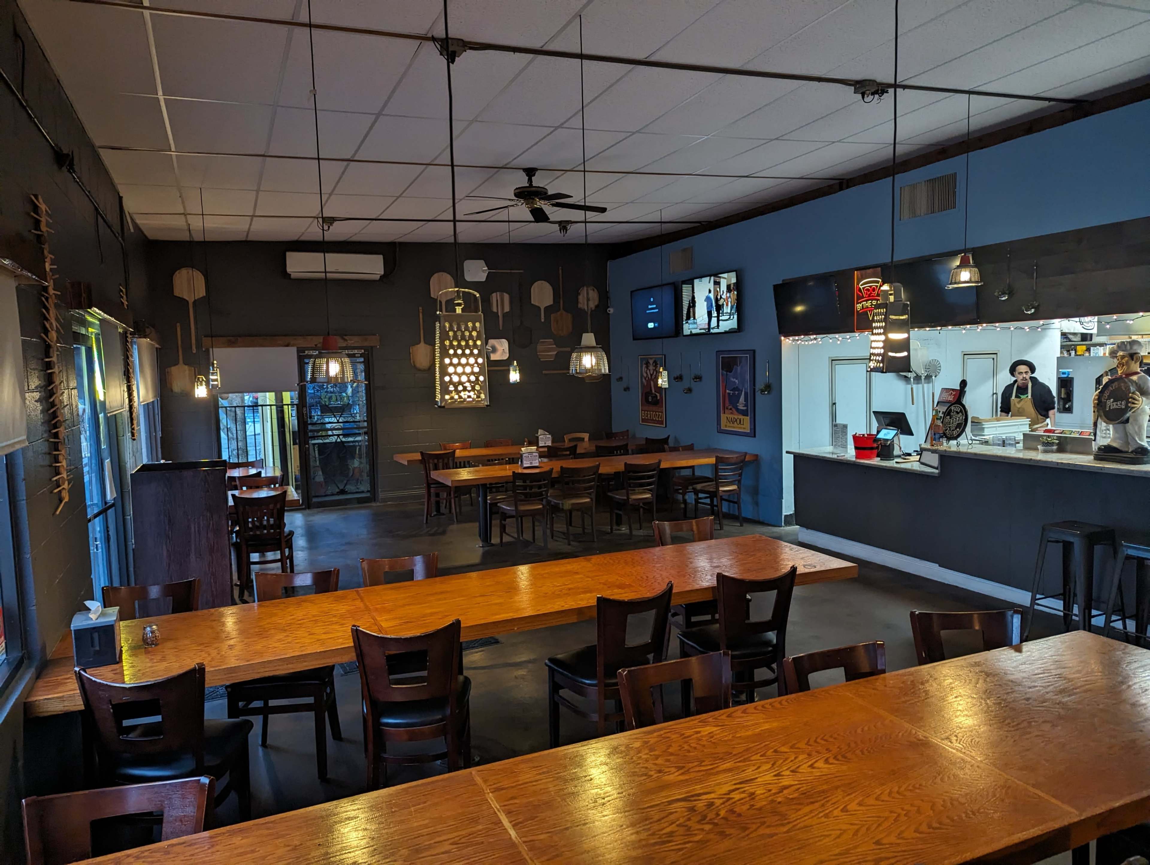 The interior of a restaurant with wooden tables, chairs, and a serving counter, featuring a kitchen area in the background.