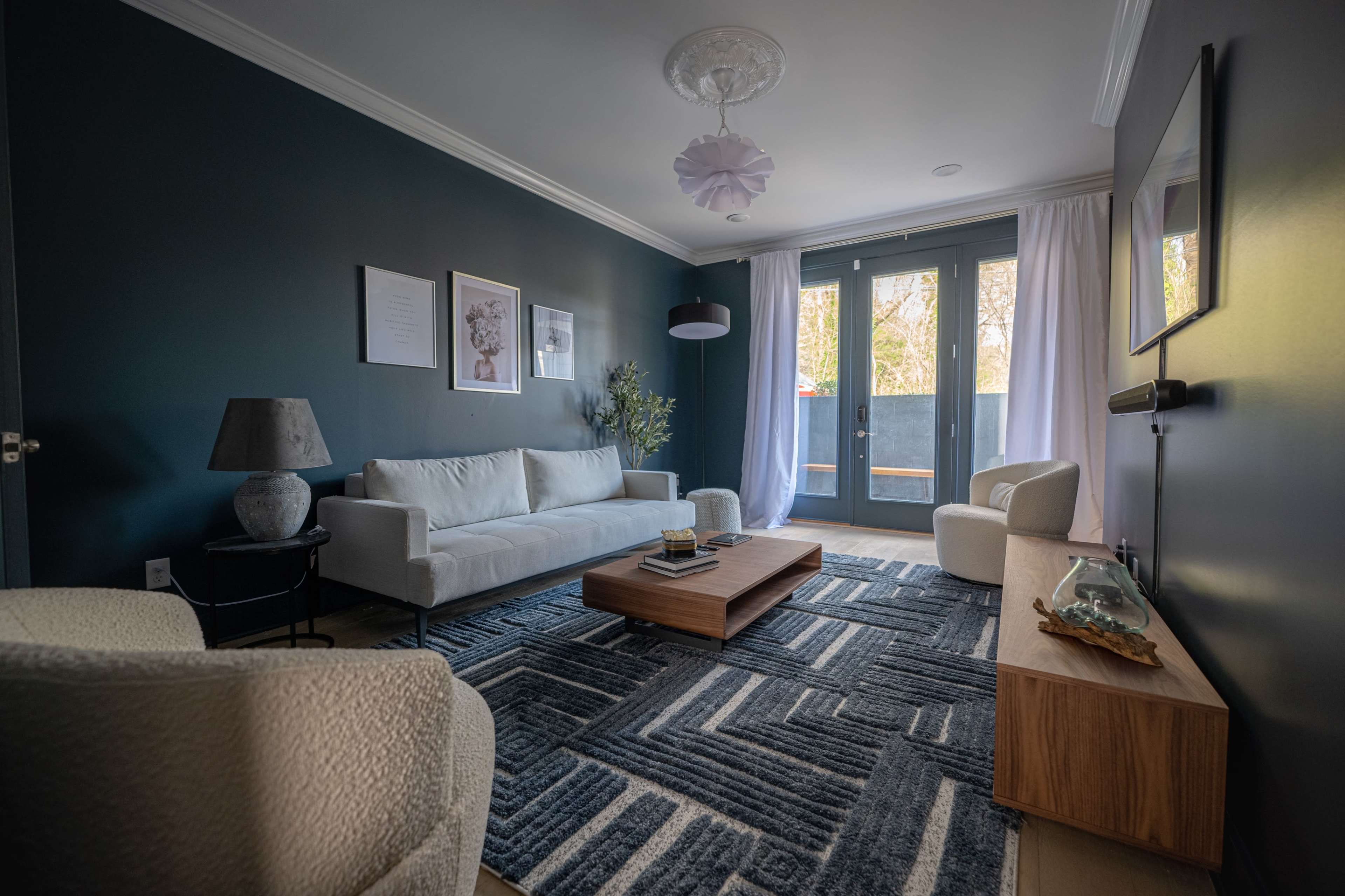 A modern living room features a light-colored sofa, a wooden coffee table, and large windows with sheer curtains, all set against dark blue walls.
