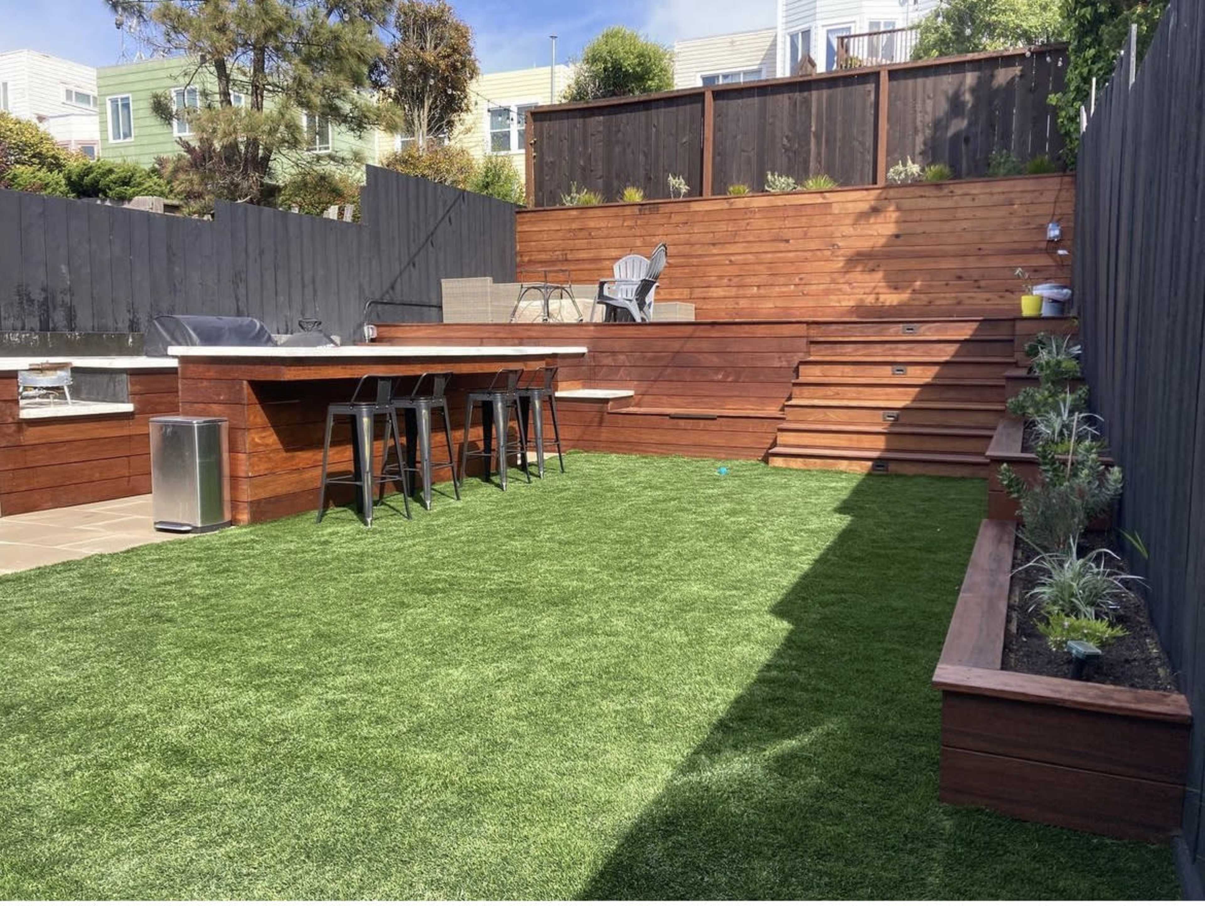 A backyard with artificial grass, wooden seating areas, a bar with stools, and steps leading to a higher level, surrounded by wooden fencing and landscaping.
