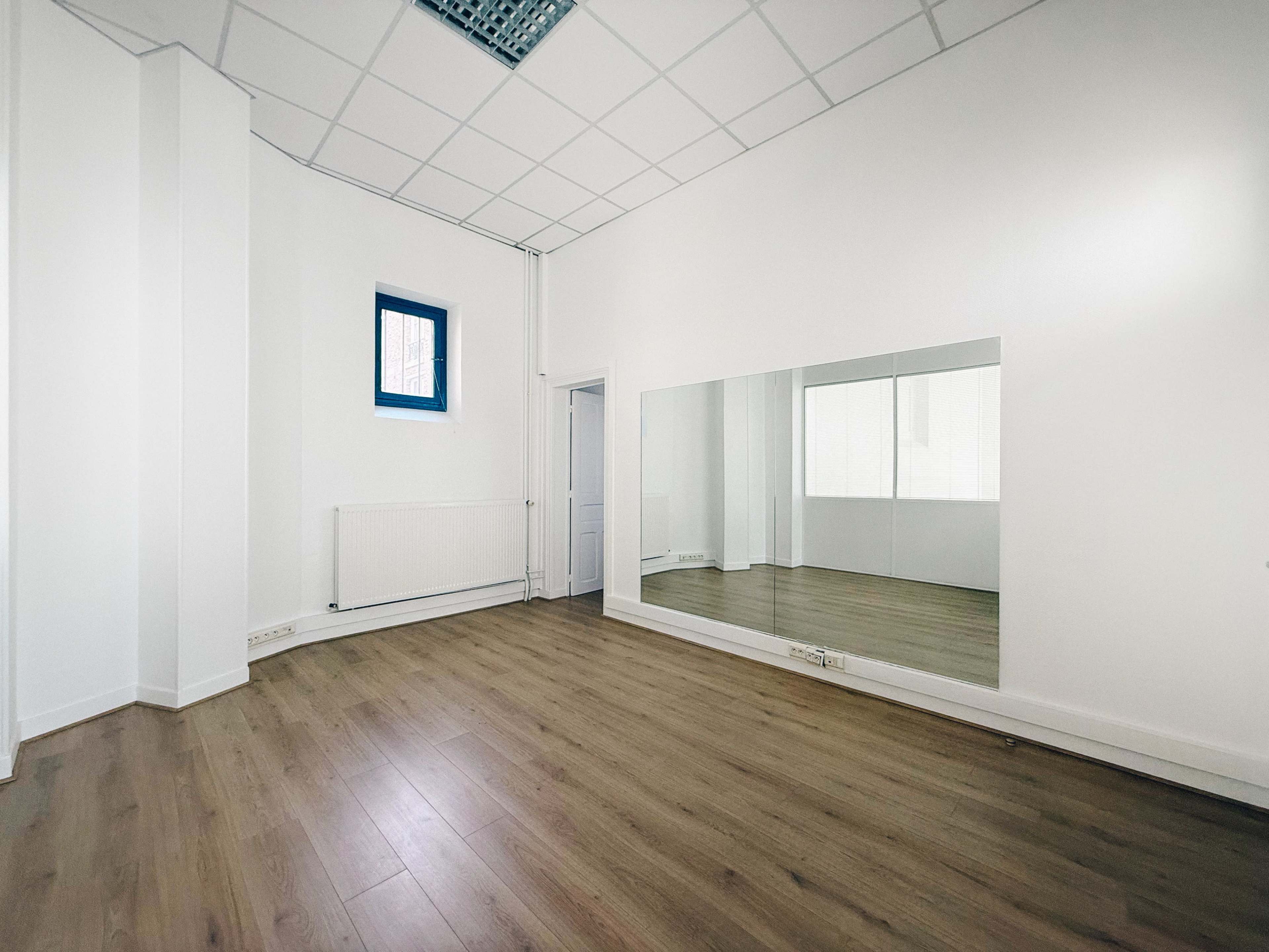 The image shows a bright, empty room with light wooden flooring and a large mirror on one wall.