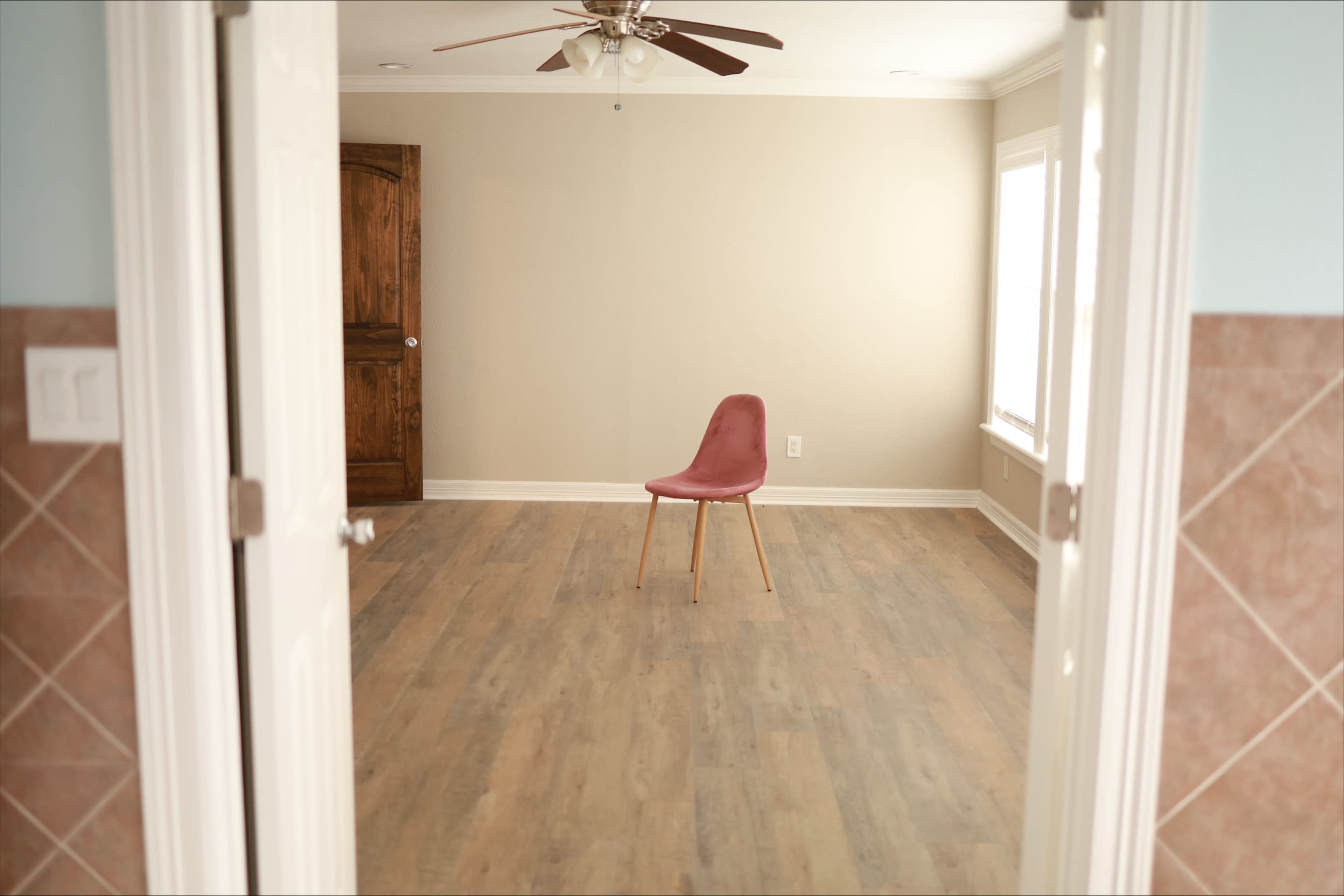 A single pink chair stands on a wooden floor in an empty room with beige walls and two windows.