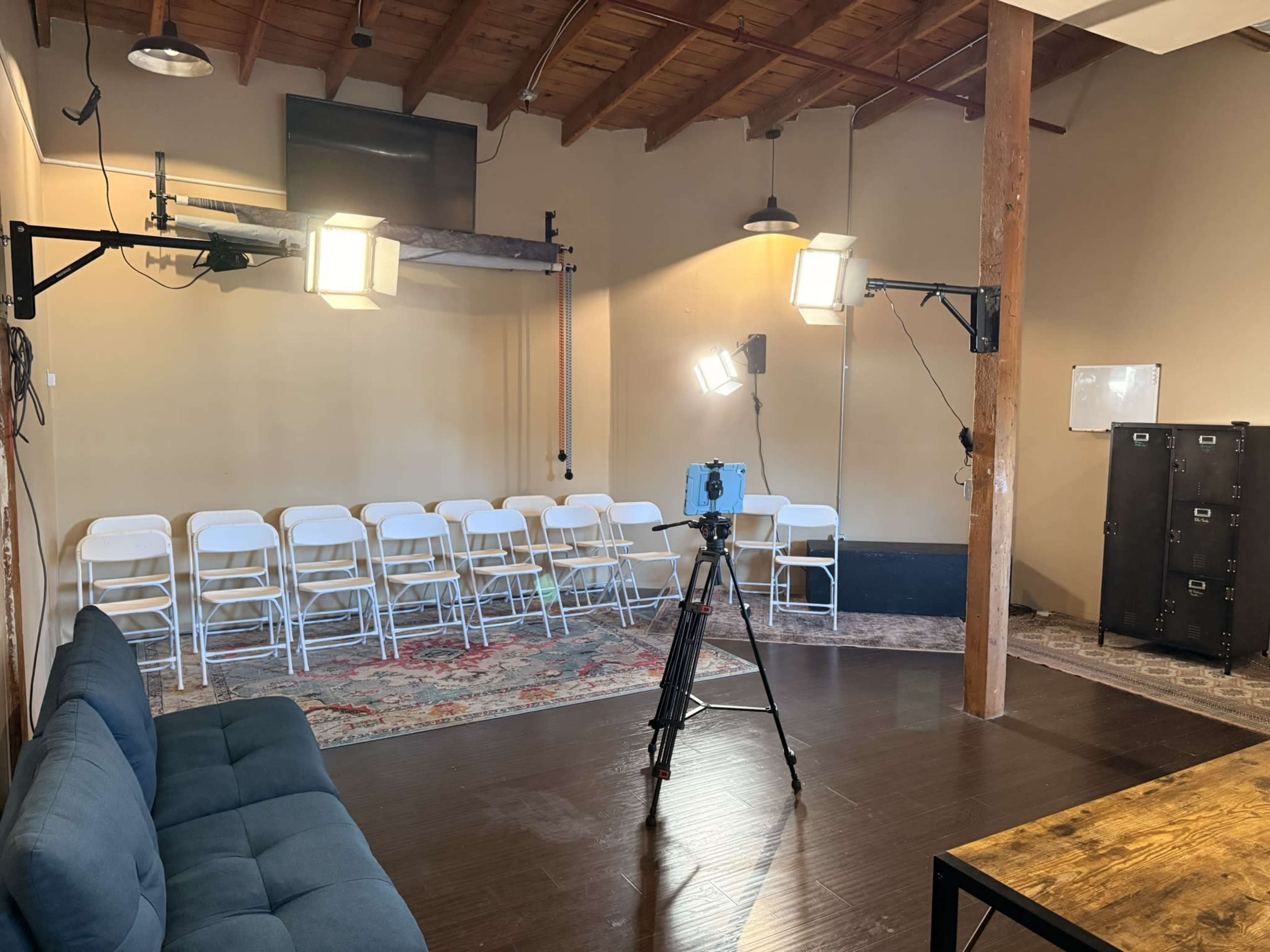 The image shows an empty room with white chairs arranged in rows, a camera on a tripod, and studio lights set up for a shoot.