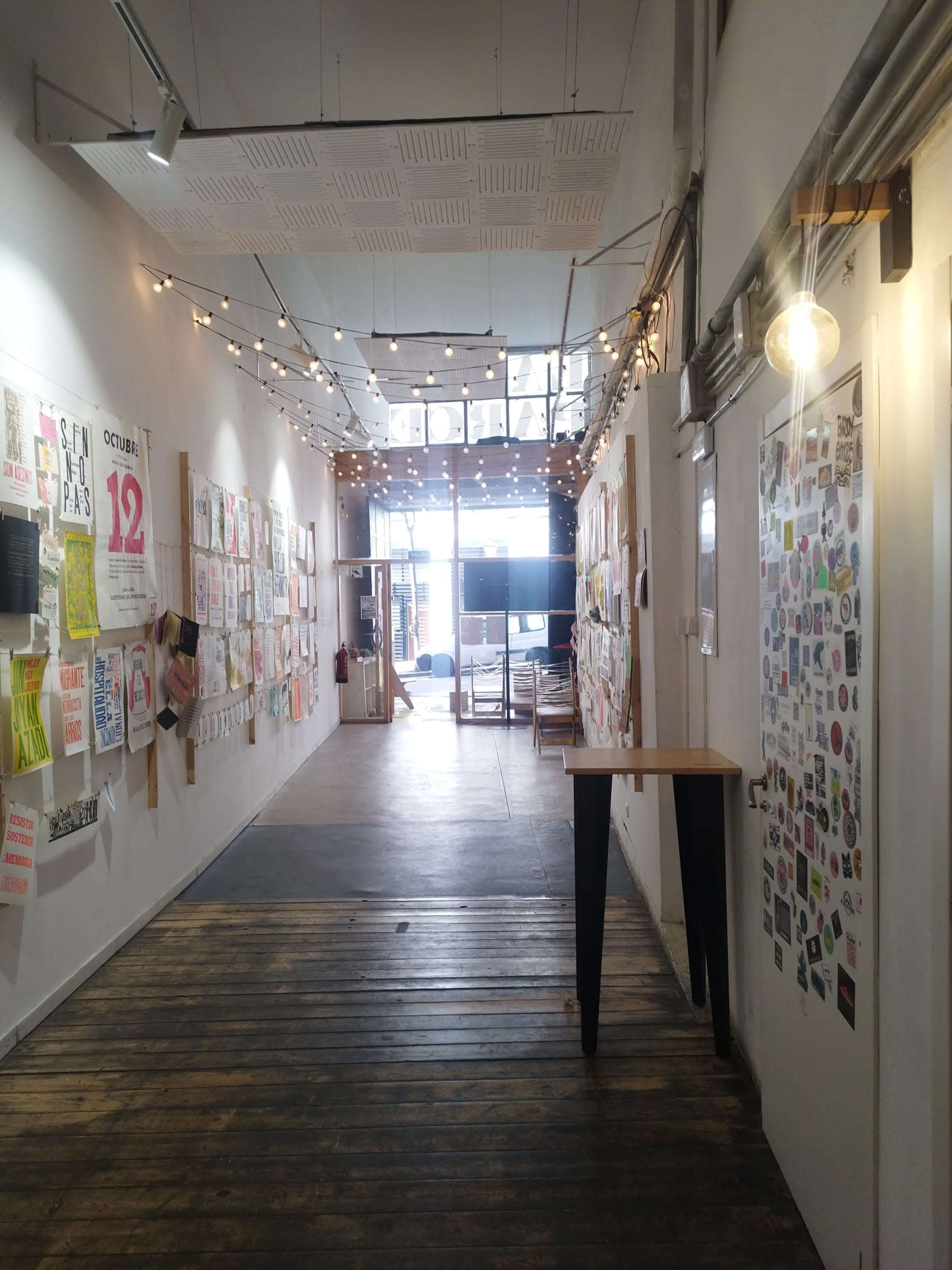 The image shows a narrow hallway adorned with colorful posters on the walls and string lights overhead, leading to an open area at the end.