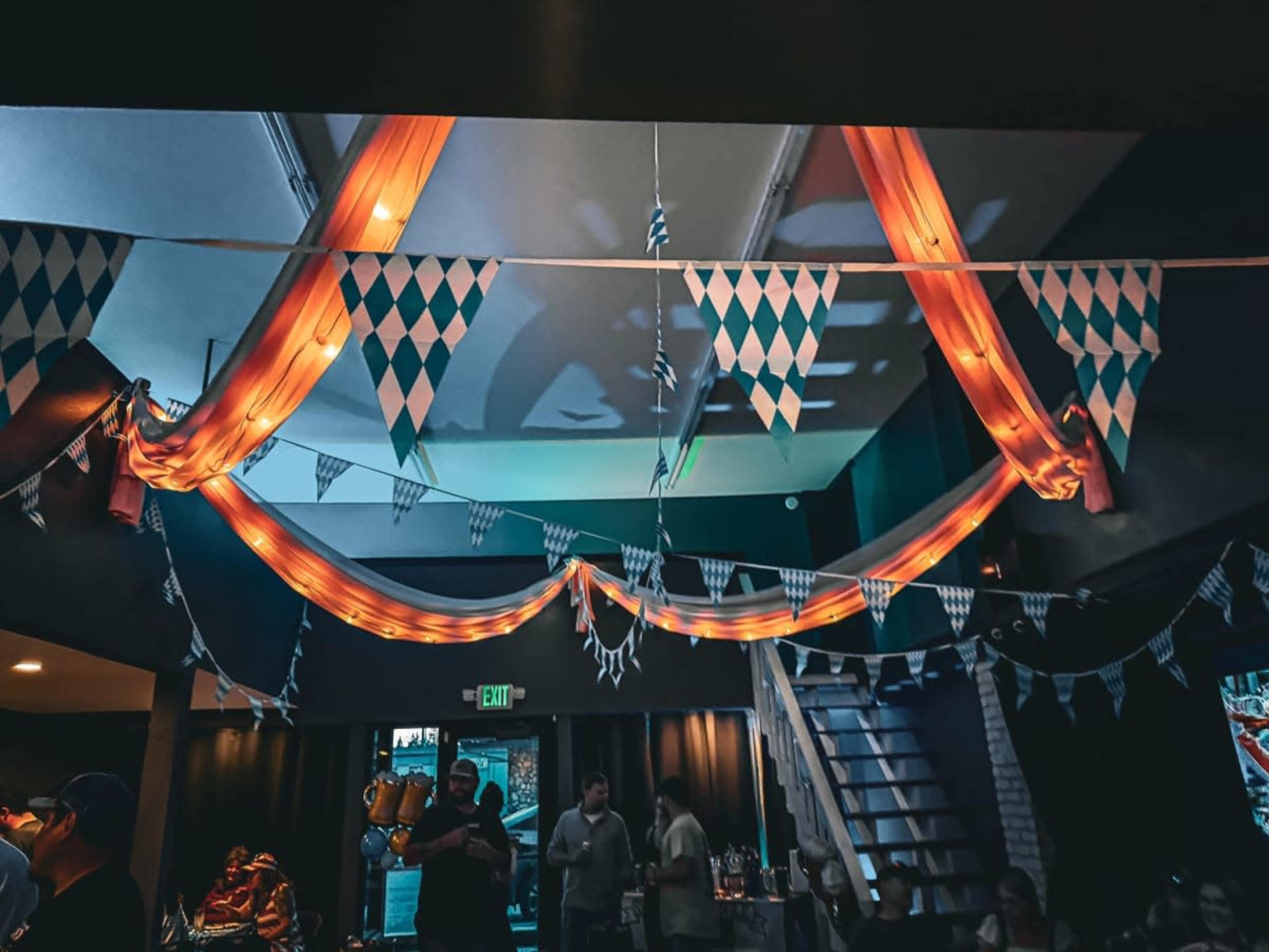 The image shows a dimly lit venue decorated with colorful bunting and draped fabric, with people socializing in the background.