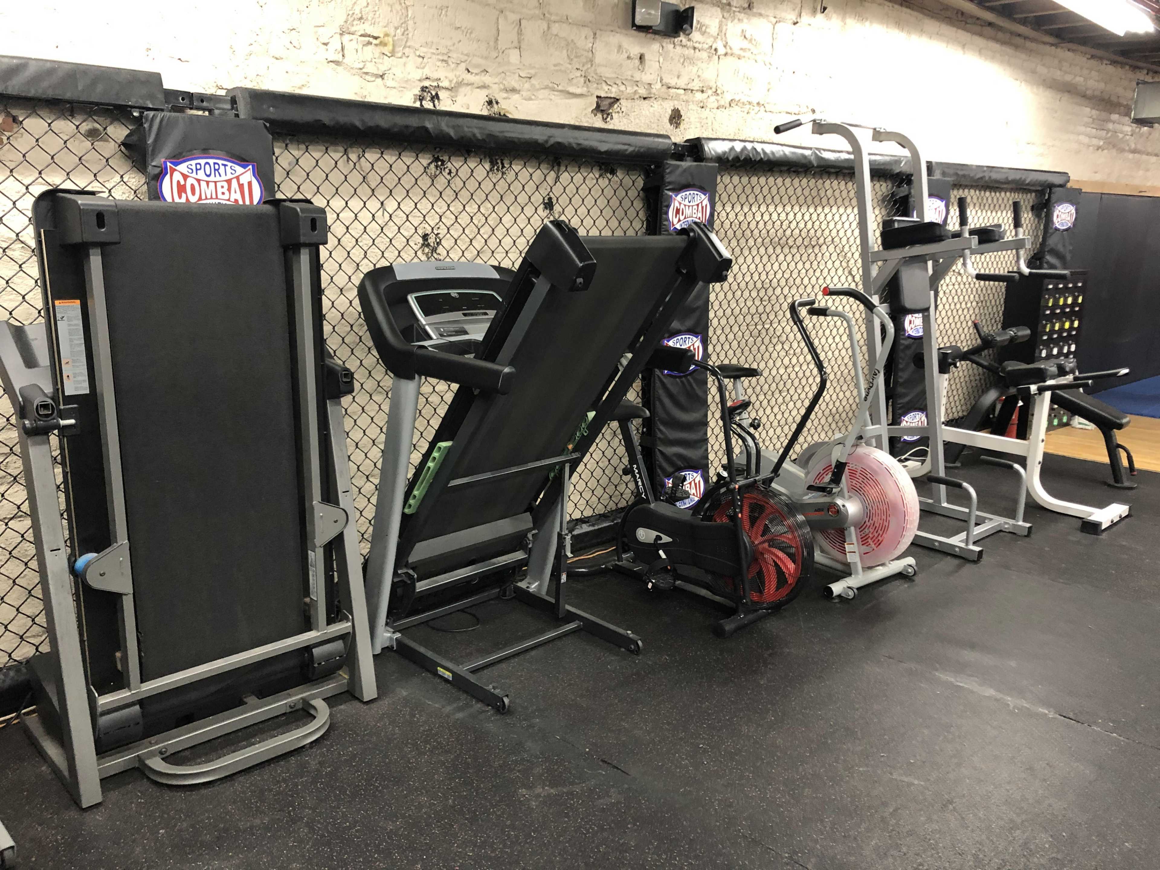 The image shows several pieces of exercise equipment lined up against a wall in a gym, including treadmills, an elliptical machine, and a weight station.