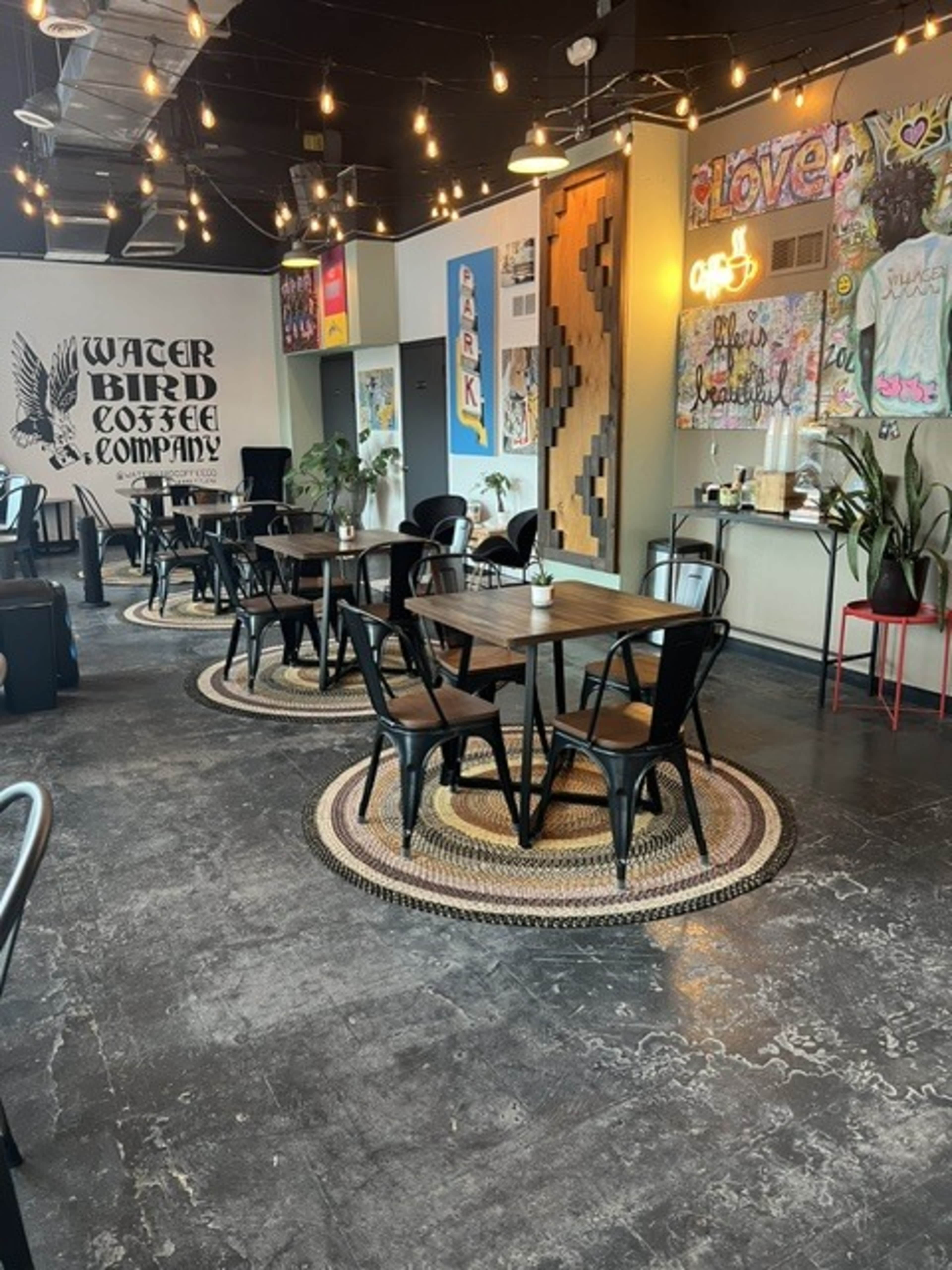 The image shows a cozy coffee shop interior with wooden tables and black chairs arranged on circular mats, adorned with colorful artwork and string lights overhead.