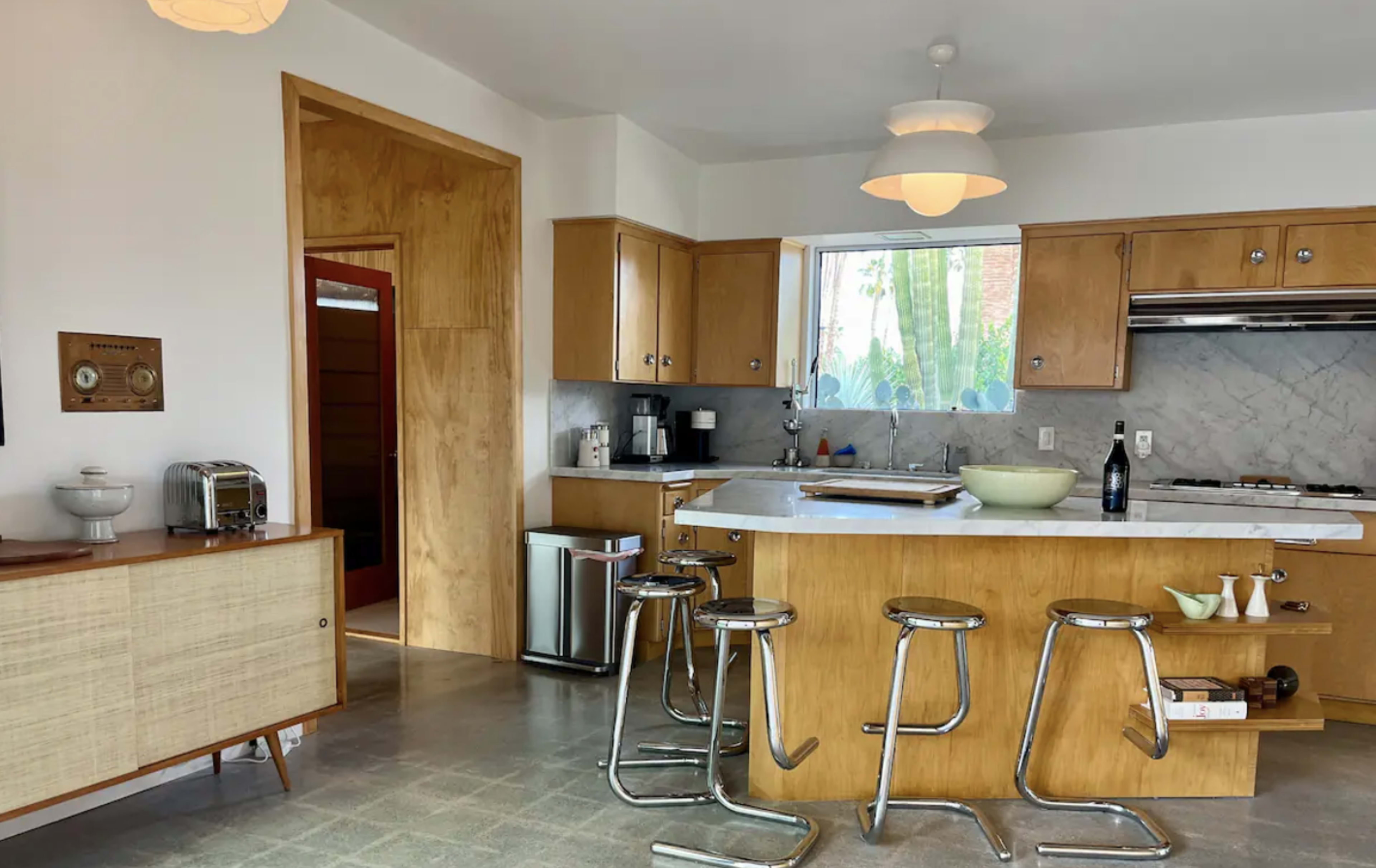 The kitchen features wooden cabinetry, a central island with bar stools, and a window overlooking a green outdoor area.