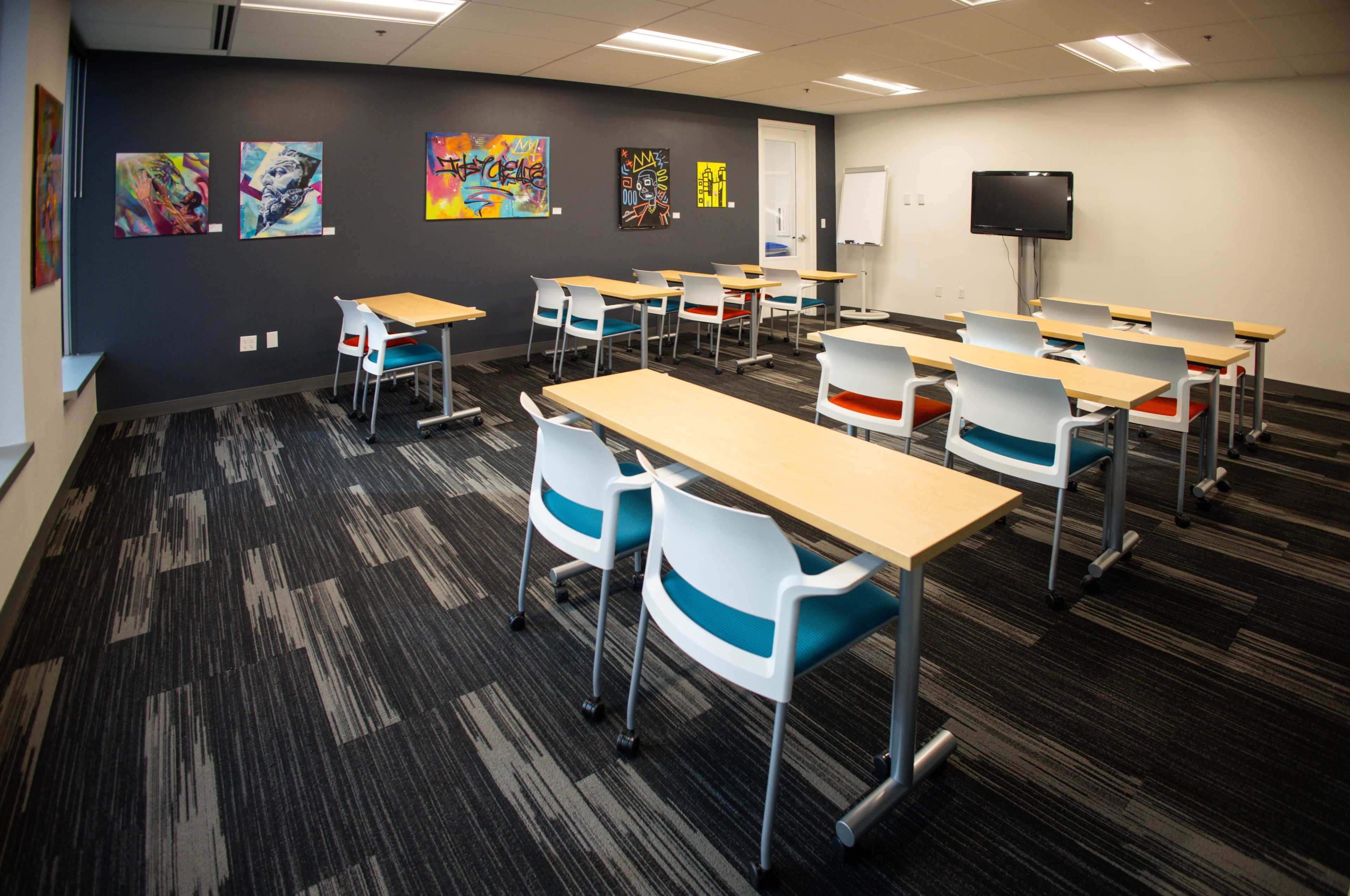 The image shows a modern classroom with several tables and chairs arranged for seating, featuring colorful wall art and a display screen at one end.