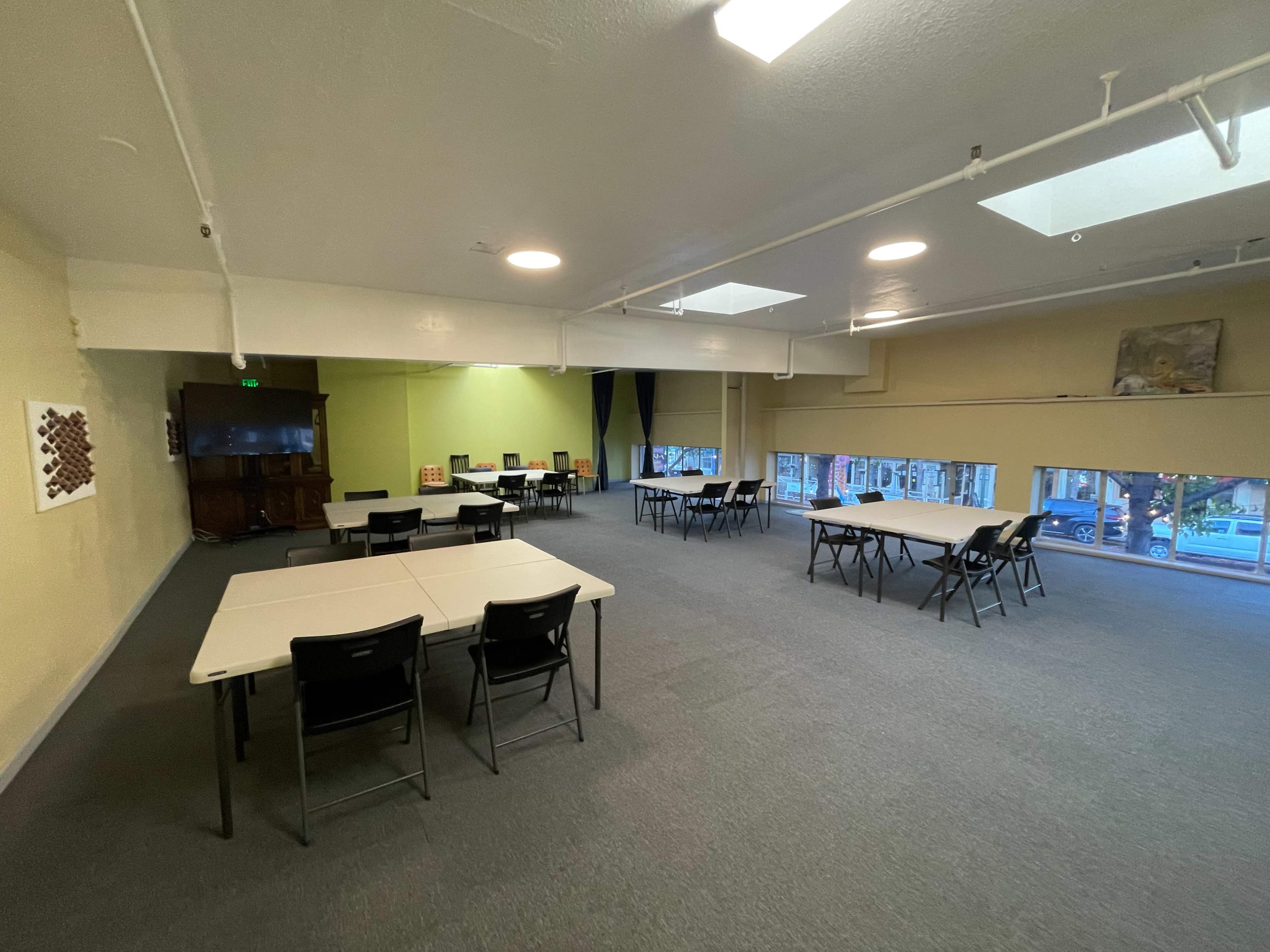 A spacious, well-lit room set up with several tables and chairs arranged for an event, with large windows providing natural light.